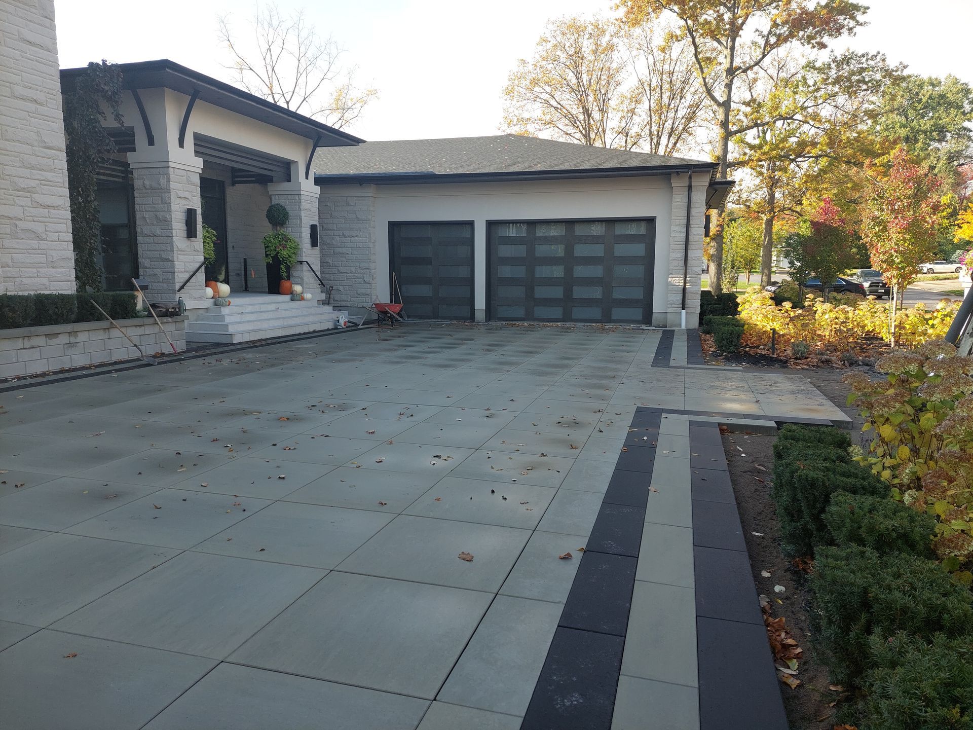 A modern home with a gray stone patio driveway leading to a two-car garage and a front porch, surrounded by fall foliage.