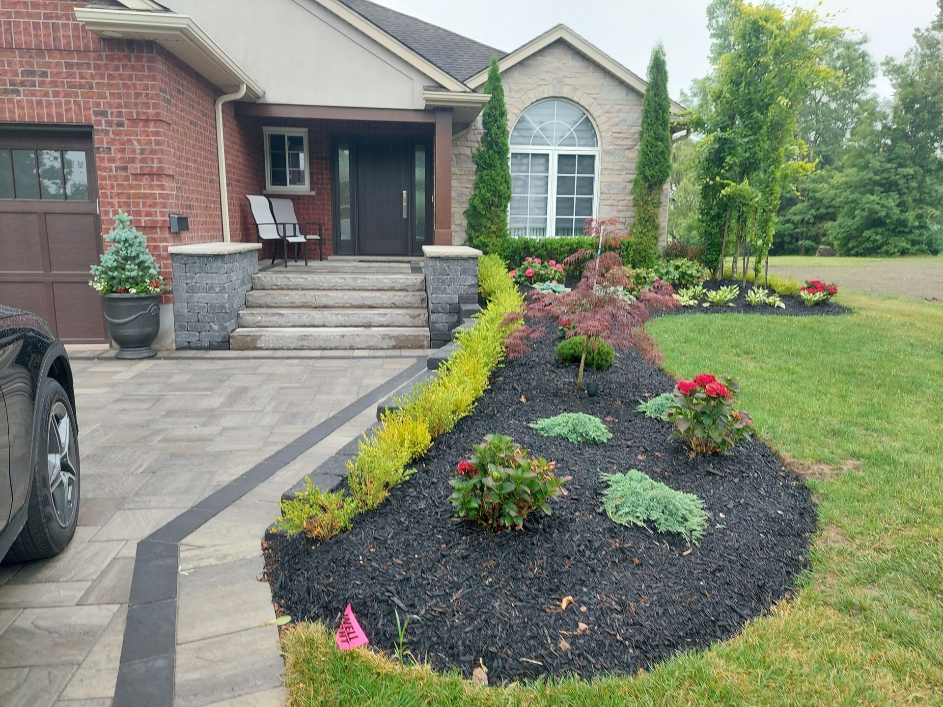 A house exterior with a paved driveway, stone steps to the front door, and a landscaped garden bed with mulch and shrubs.