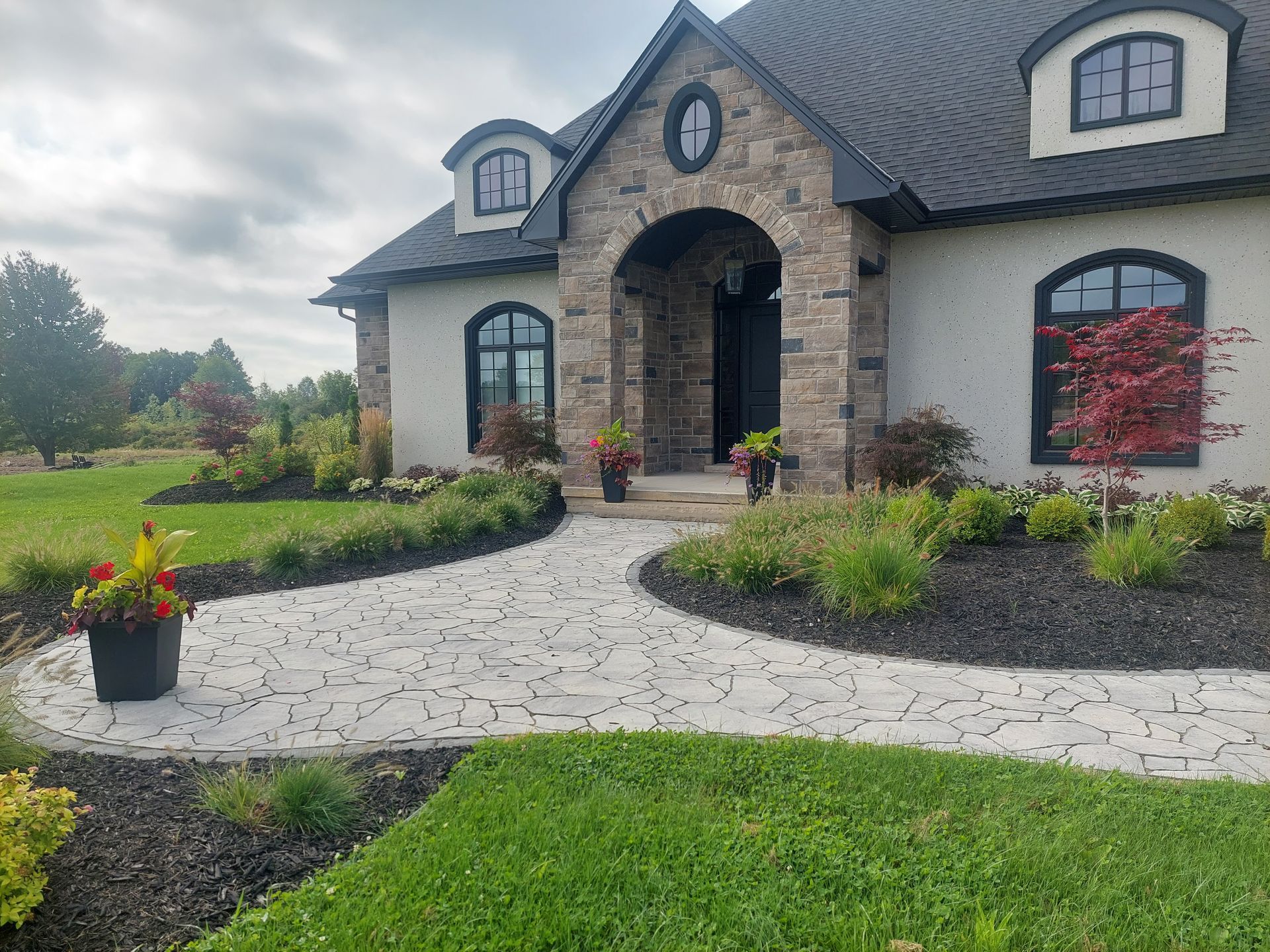 A stone-veneer house with a curved flagstone walkway, landscaped garden beds, and dark window frames under a cloudy sky.