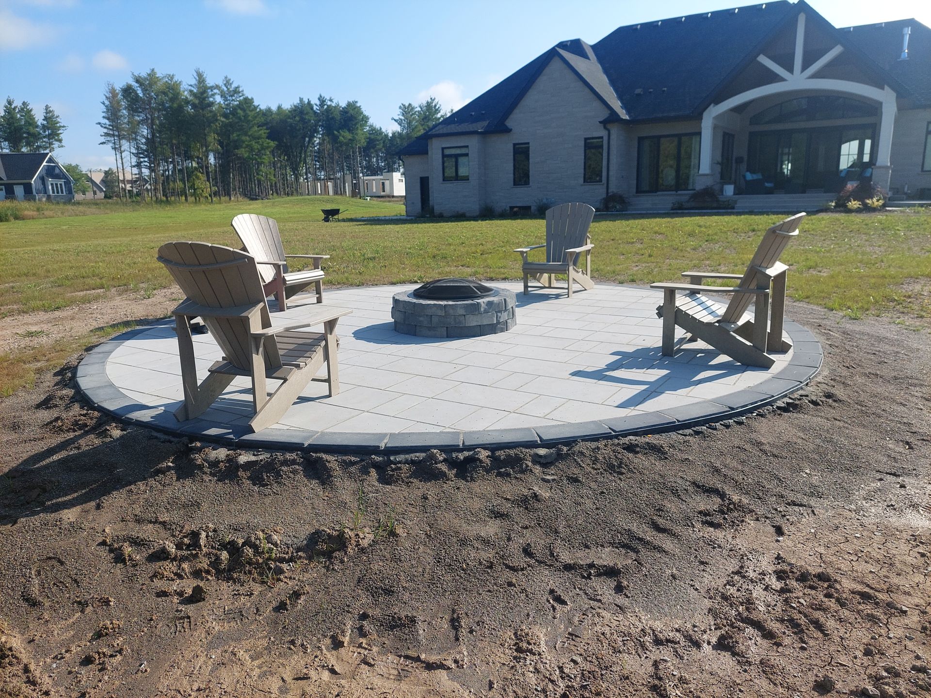 A circular paver patio with a stone fire pit and four chairs in a grassy backyard next to a residential home.