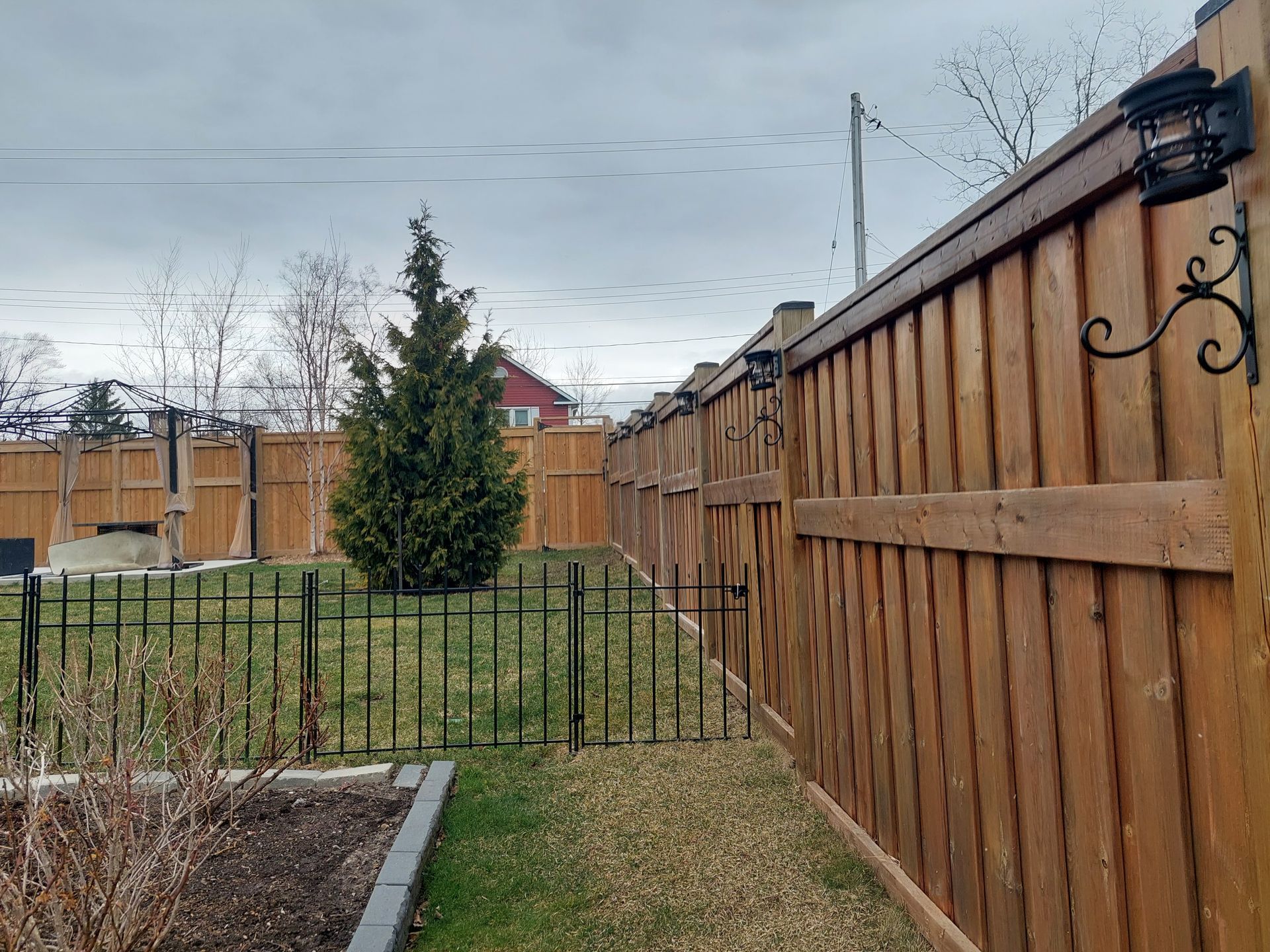 A backyard view with a wooden fence featuring mounted lights and a black metal garden fence enclosing a patch of lawn.