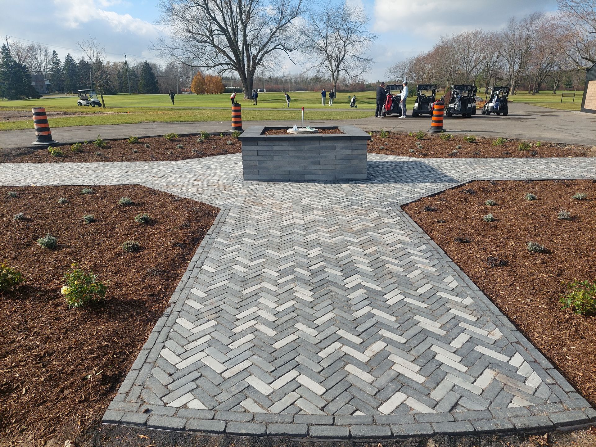 A herringbone-patterned stone walkway leads to a square fire pit feature in a golf course landscape with scattered golfers.