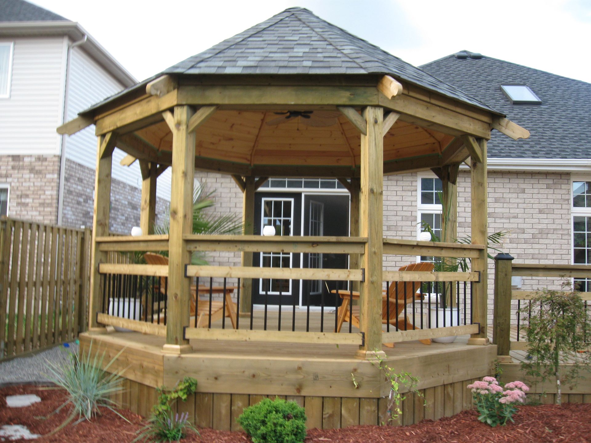 A wooden, hexagonal gazebo sits on a deck in a backyard, featuring a dark shingled roof and black metal railing accents.
