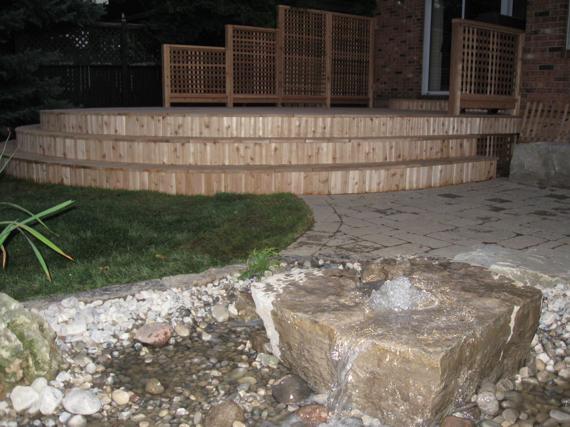 A wooden tiered deck with privacy lattice screens overlooks a stone water feature in a backyard.