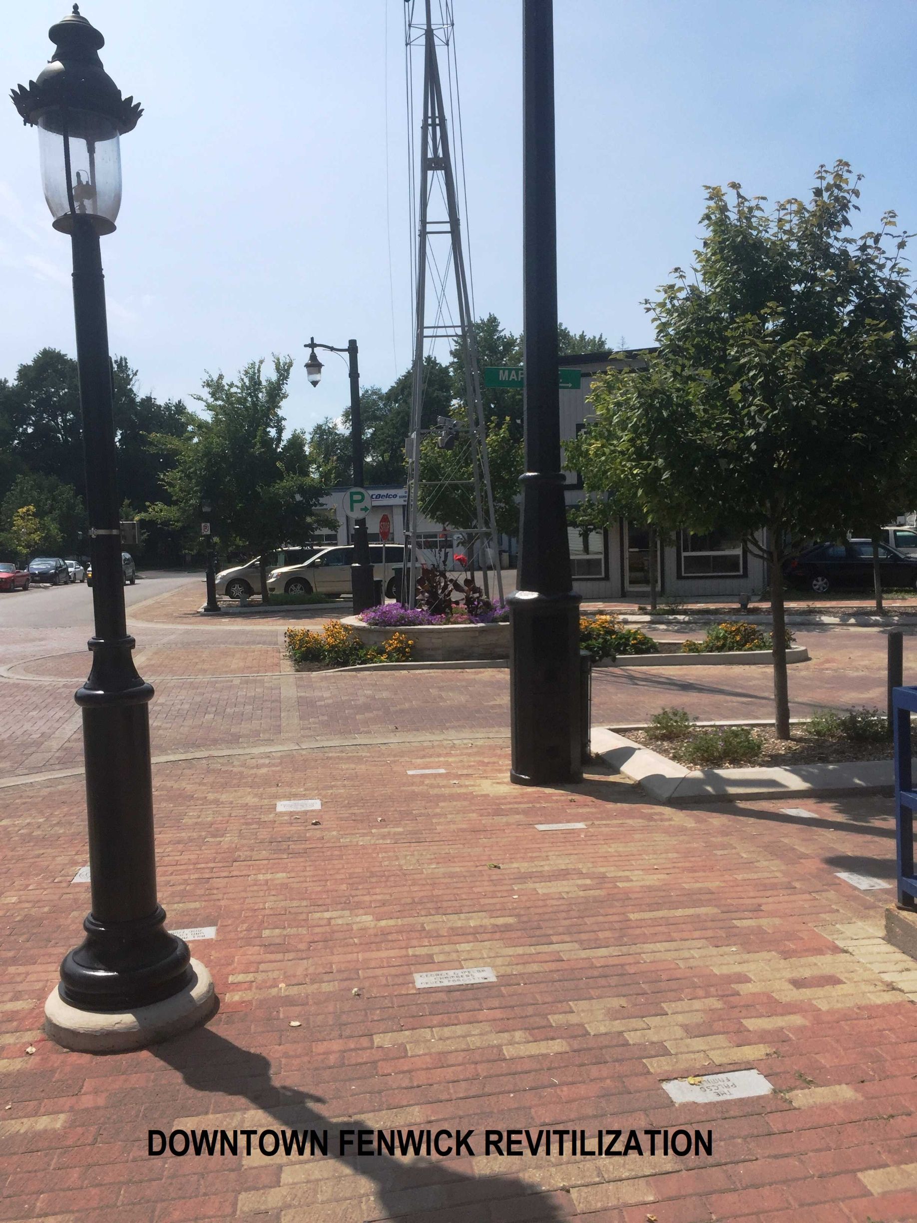A brick pedestrian area in downtown Fenwick featuring decorative lamp posts, a windmill sculpture, and trees.