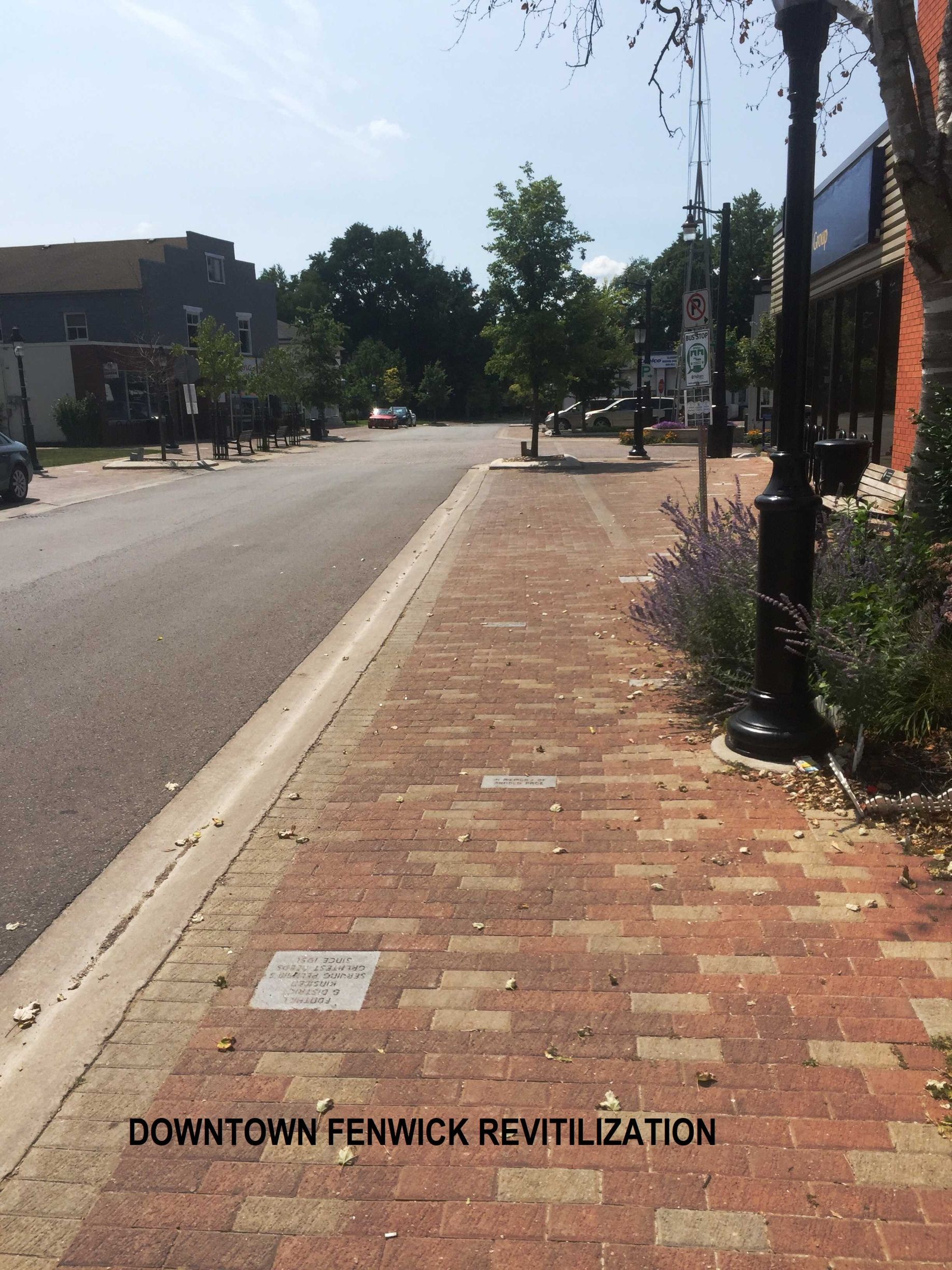 A sidewalk paved with red bricks alongside a street in a downtown area, with trees, a lamppost, and a revitalization sign.