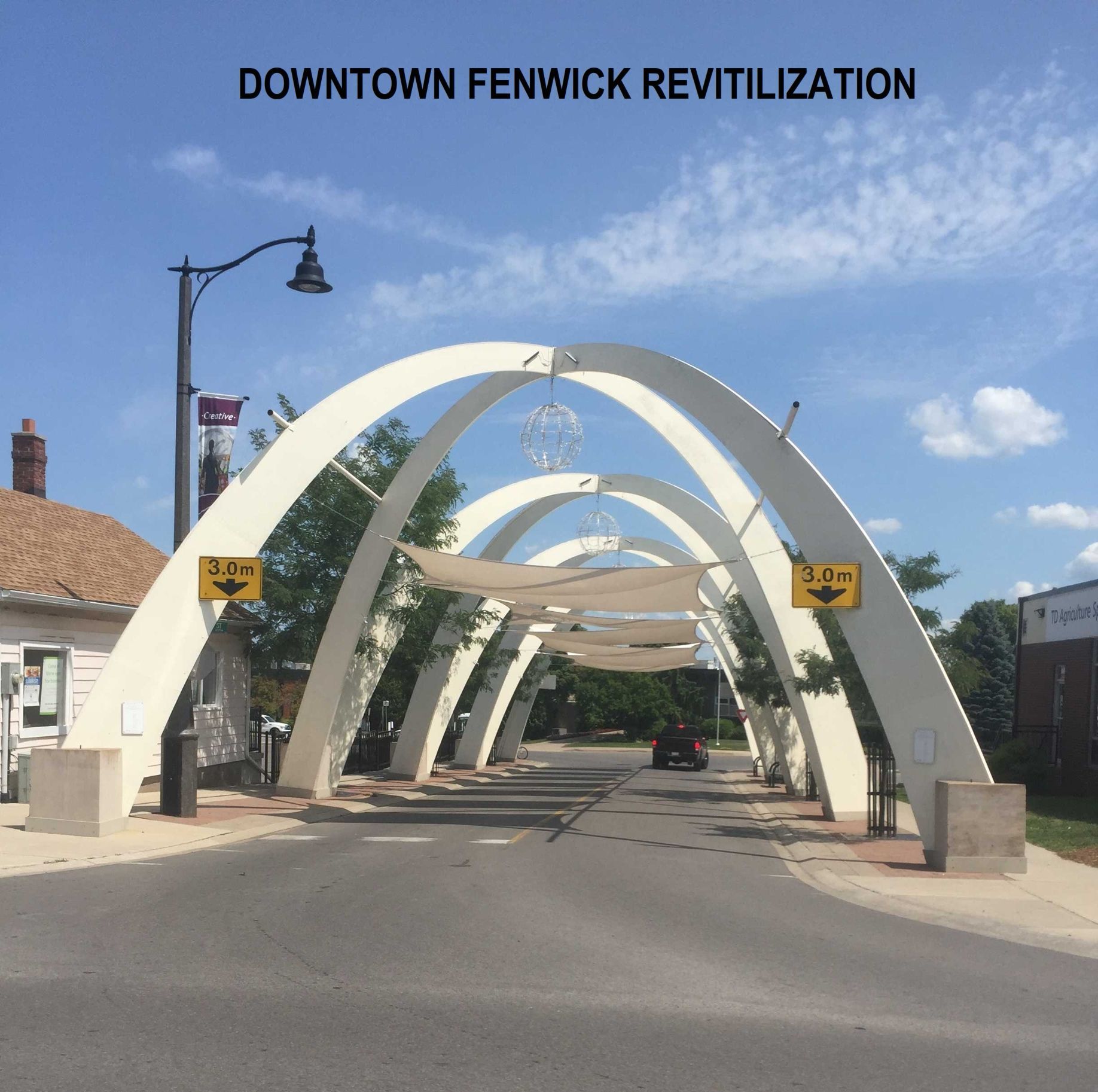 White archways span a paved street in downtown Fenwick under a bright blue sky, with yellow height restriction signs.