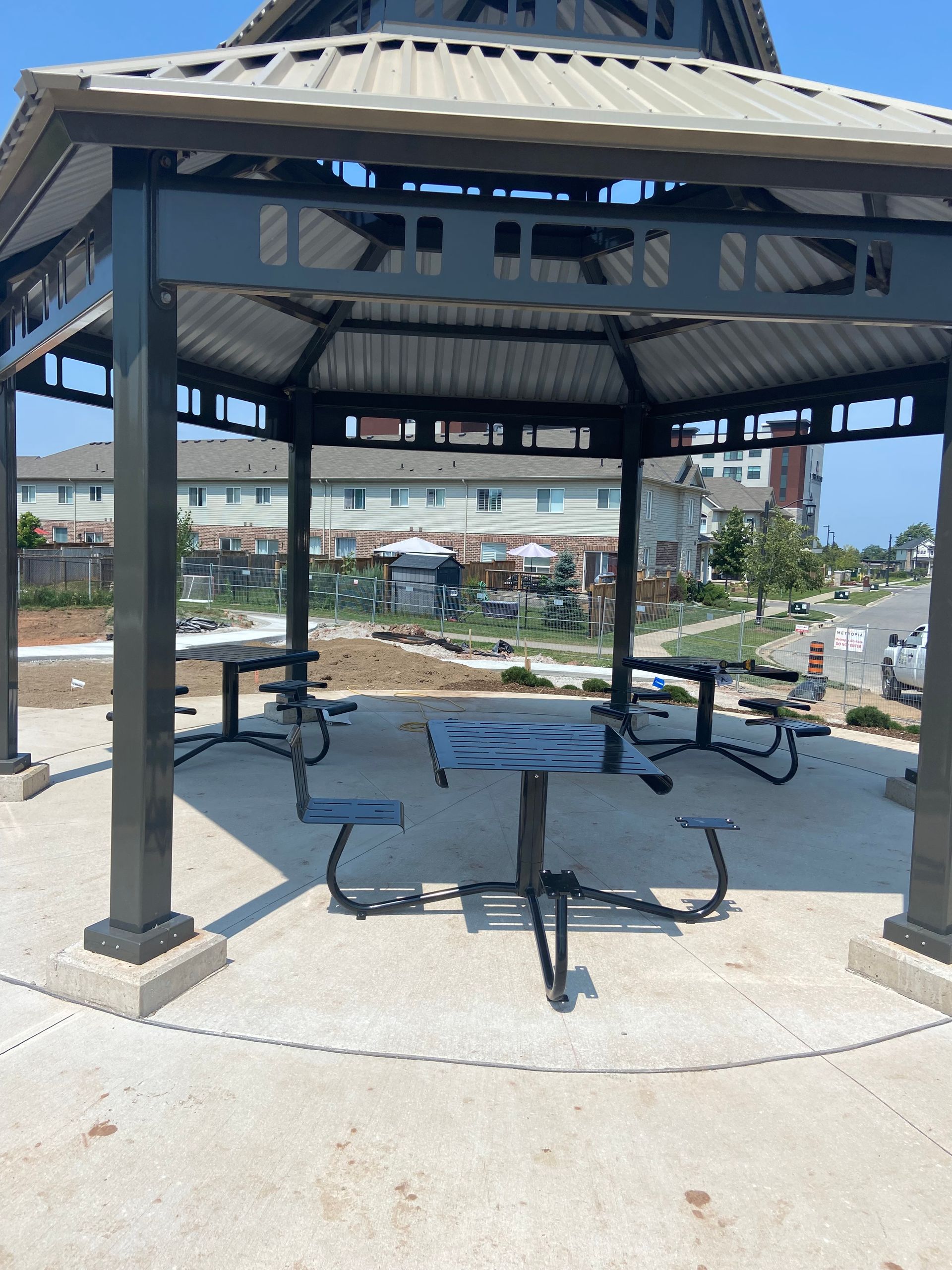 A black metal gazebo structure with built-in, matching tables and seats on a concrete pad outdoors on a sunny day.