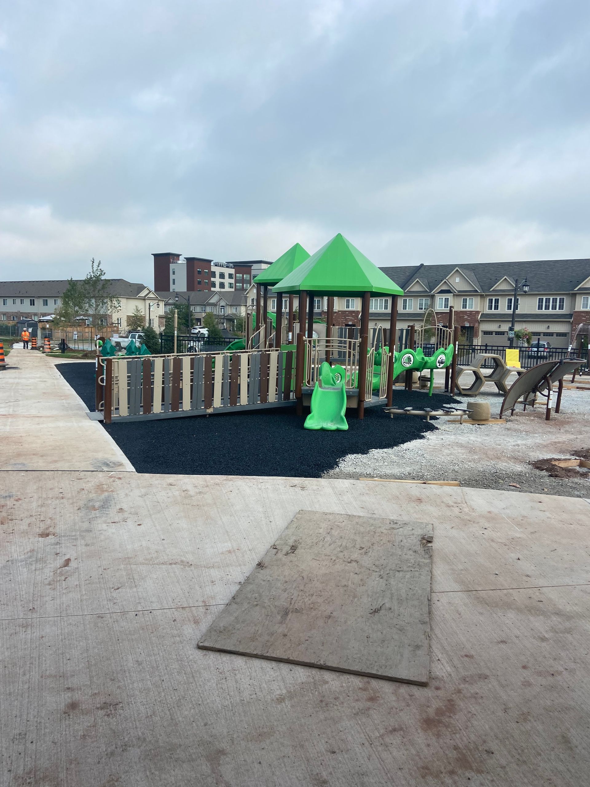 A playground with green-roofed structures and a black rubberized play surface, situated in front of residential townhomes.