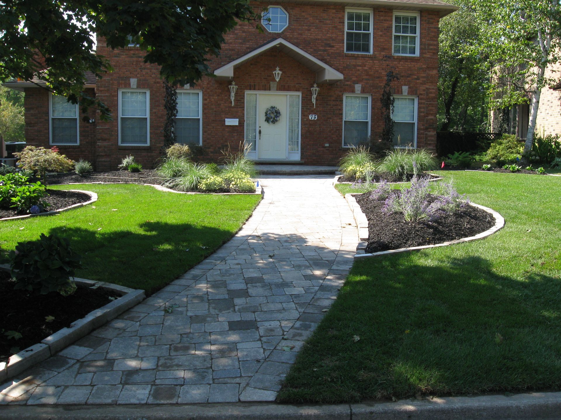 A brick house with a stone walkway leading to the front door, surrounded by a green lawn and landscaped flower beds.