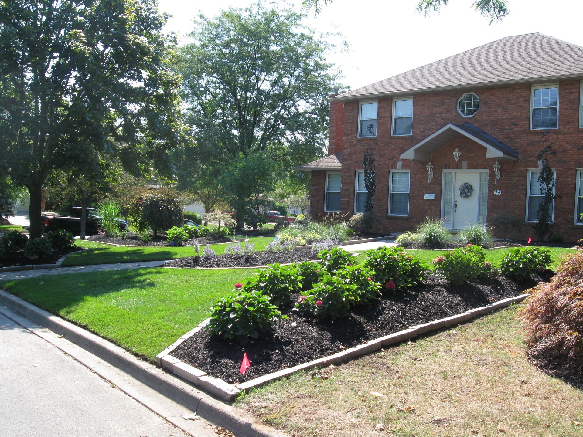 A two-story brick house with a front garden featuring a landscaped bed of shrubs and mulch, viewed from the sidewalk.
