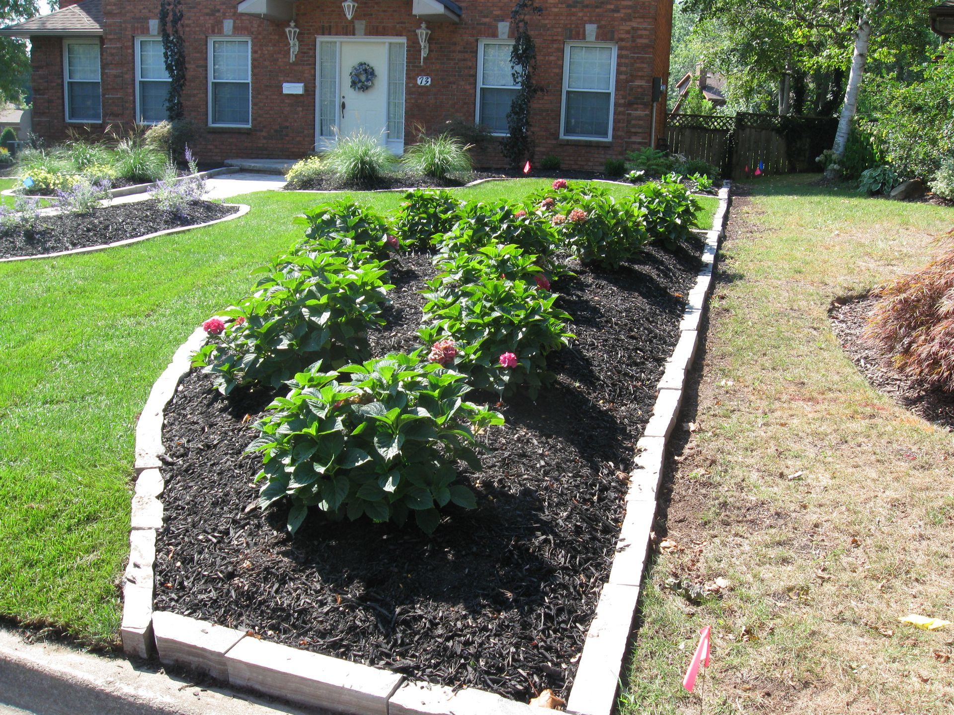 A brick house with a landscaped garden bed containing small shrubs in dark mulch, bordered by stone pavers on a green lawn.