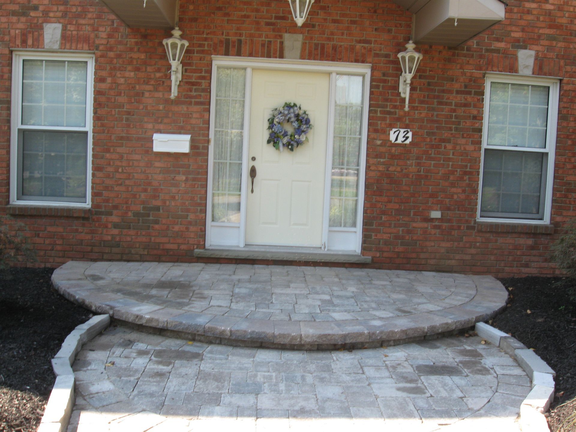 Front view of a brick house entryway featuring stone-paver steps and a patio leading to a white door with a wreath.