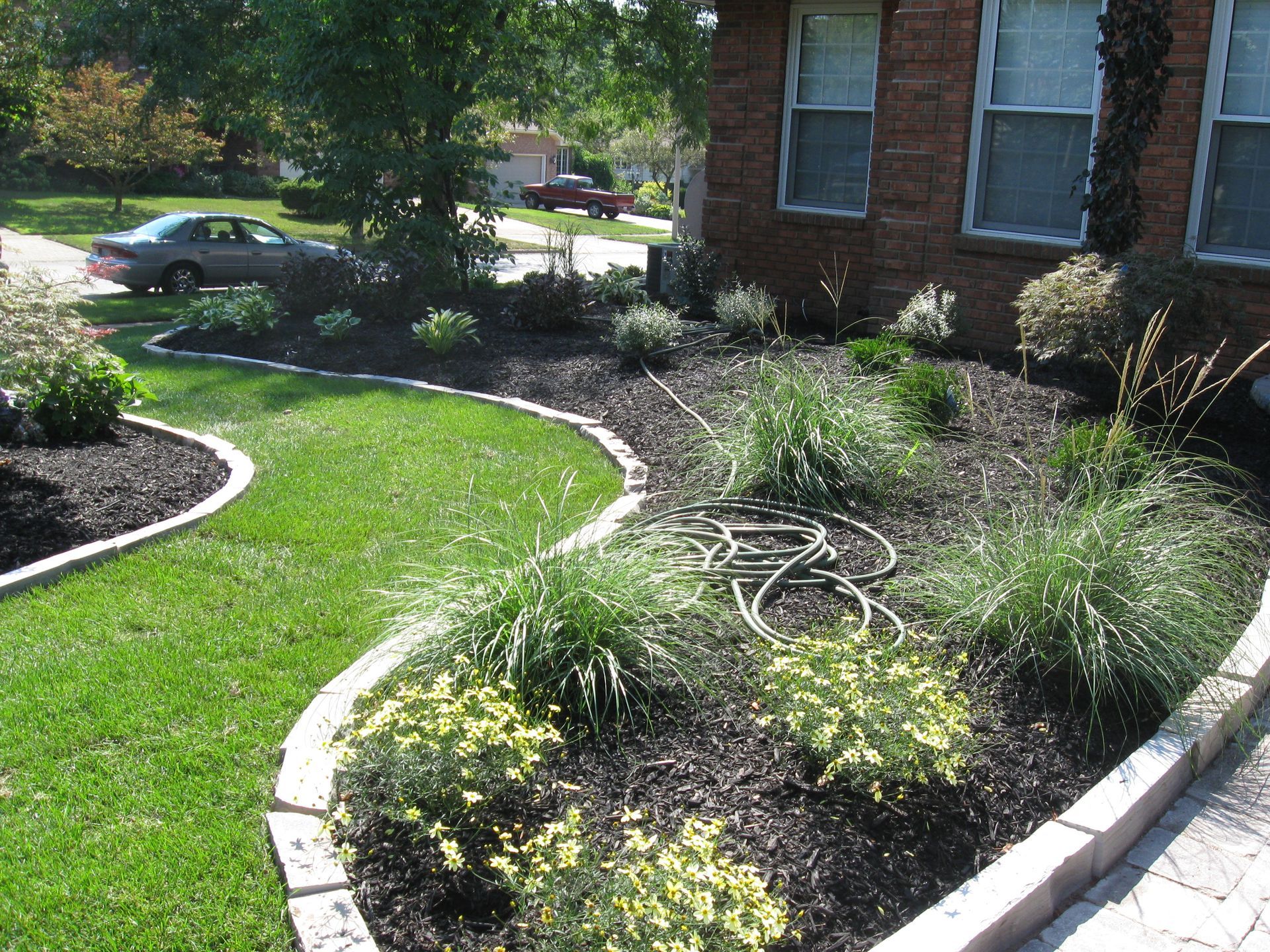 A landscaped yard with a curved stone-edged garden bed, mulch, ornamental grasses, shrubs, and a brick house exterior.