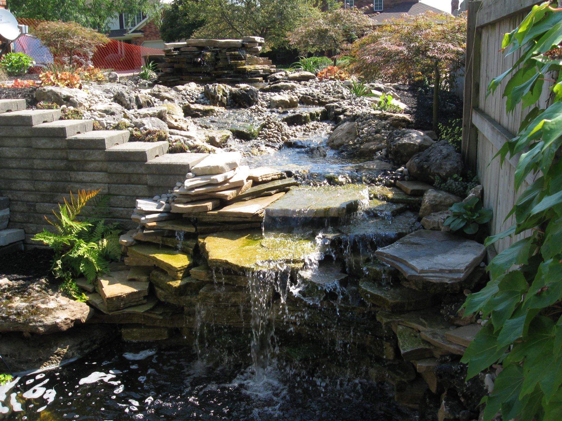 A tiered stone waterfall flows into a dark pond next to a stone retaining wall and a fence with climbing green ivy.