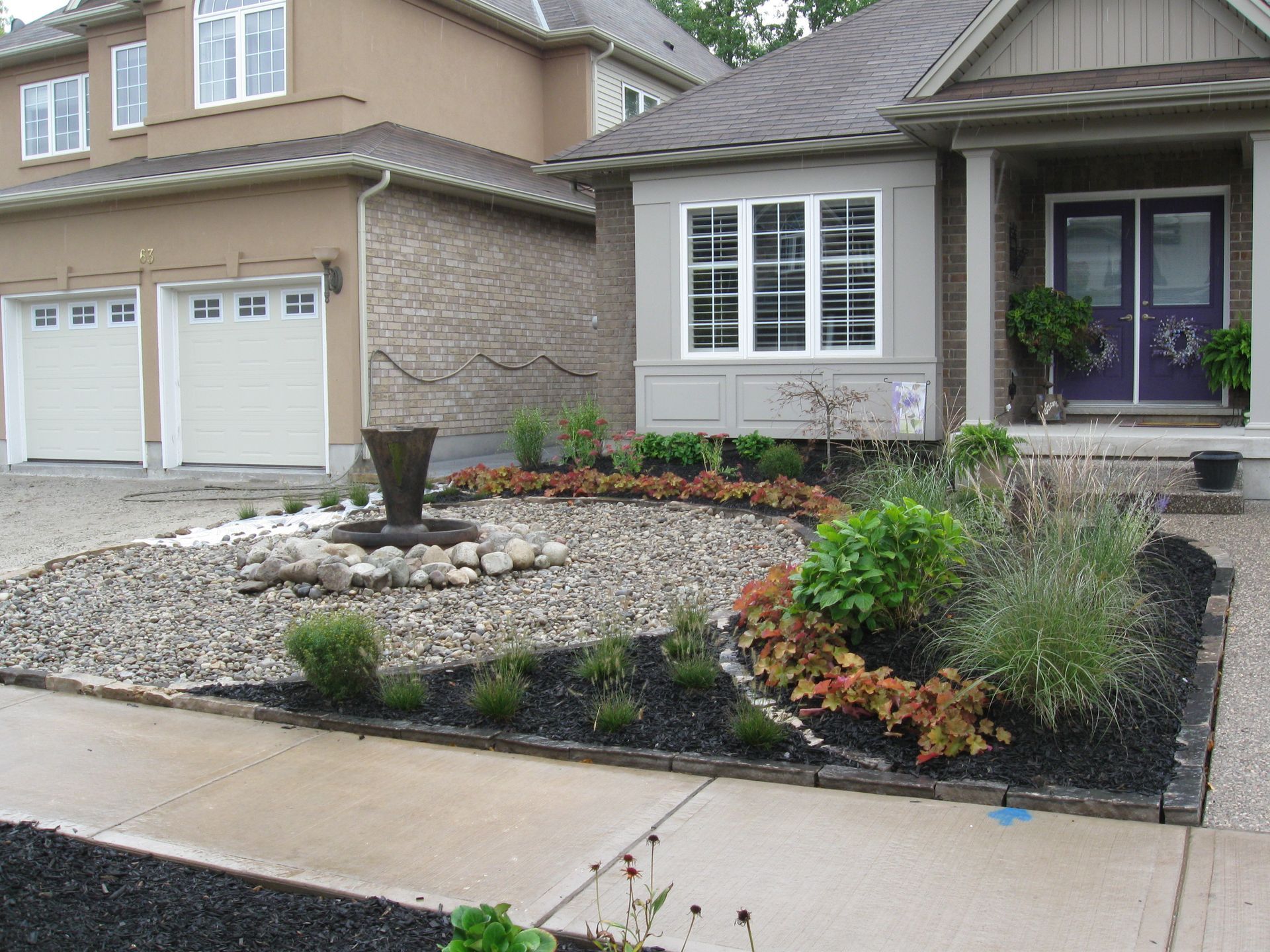 A front yard garden features a central stone fountain surrounded by gravel, black mulch beds, ornamental grasses, and shrubs.