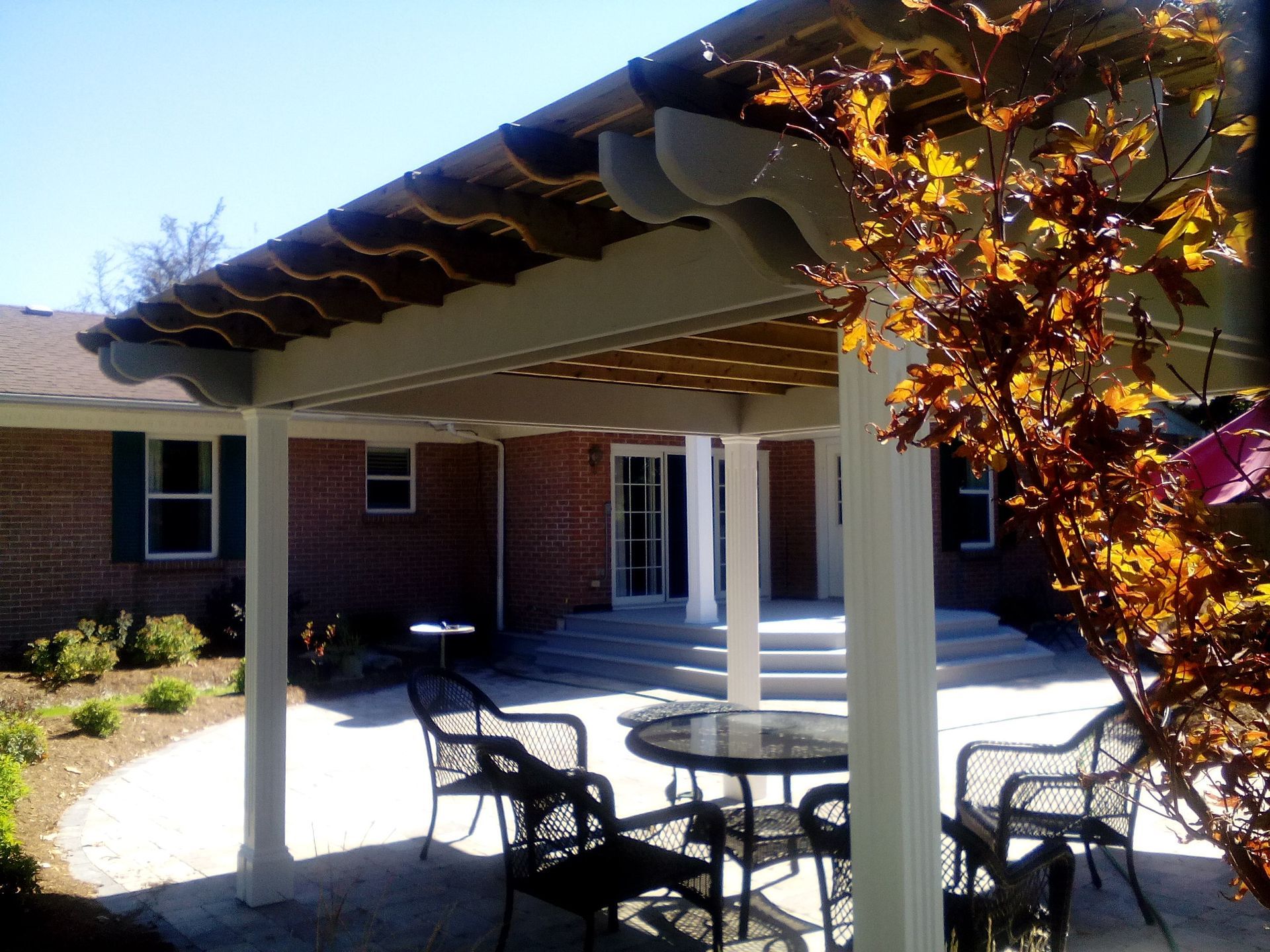 A patio with a pergola, a table, and chairs sits outside a brick house, partially obscured by autumn leaves.