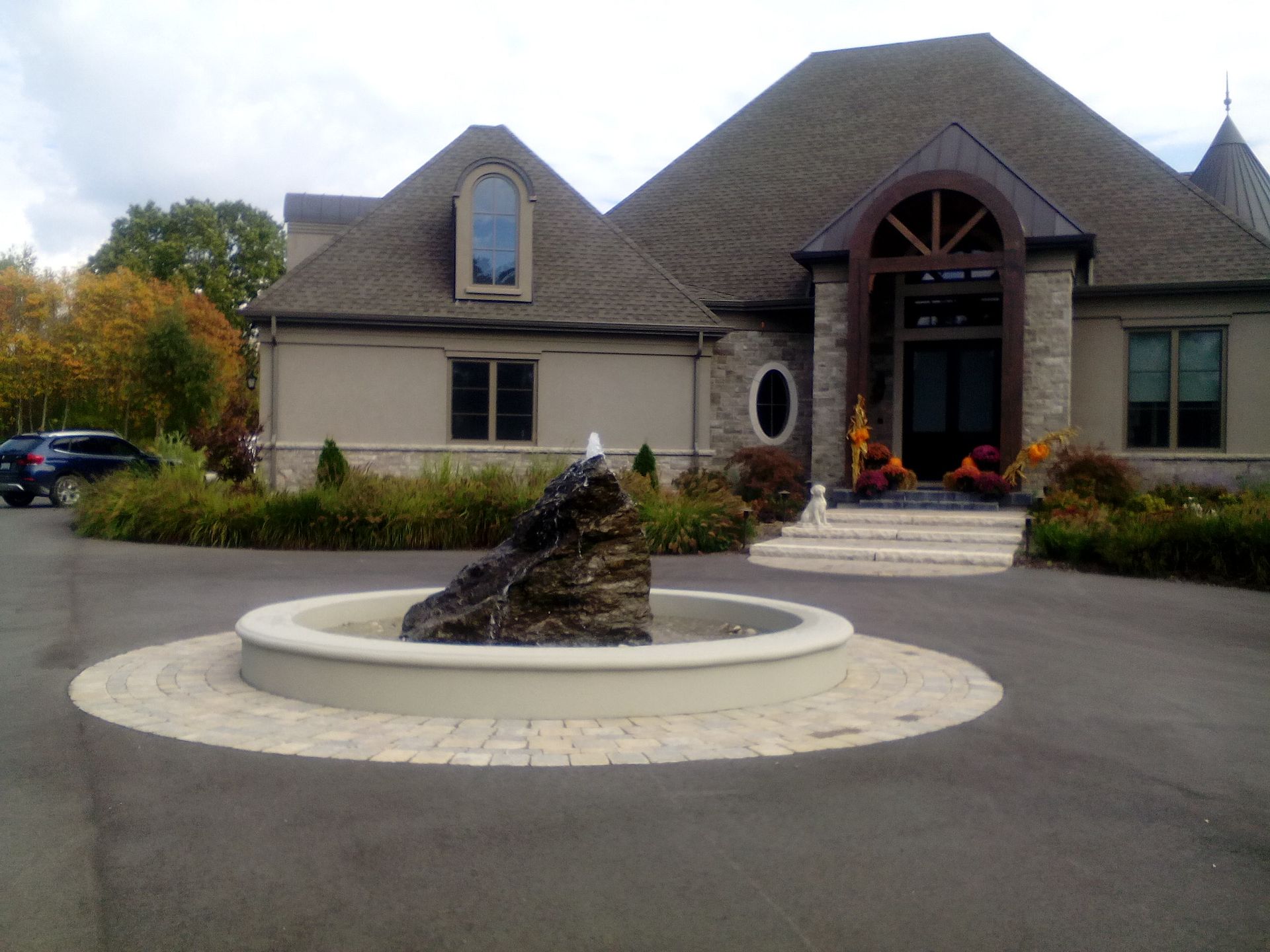 A large house with a stone entryway and a circular stone fountain in the center of a paved driveway.