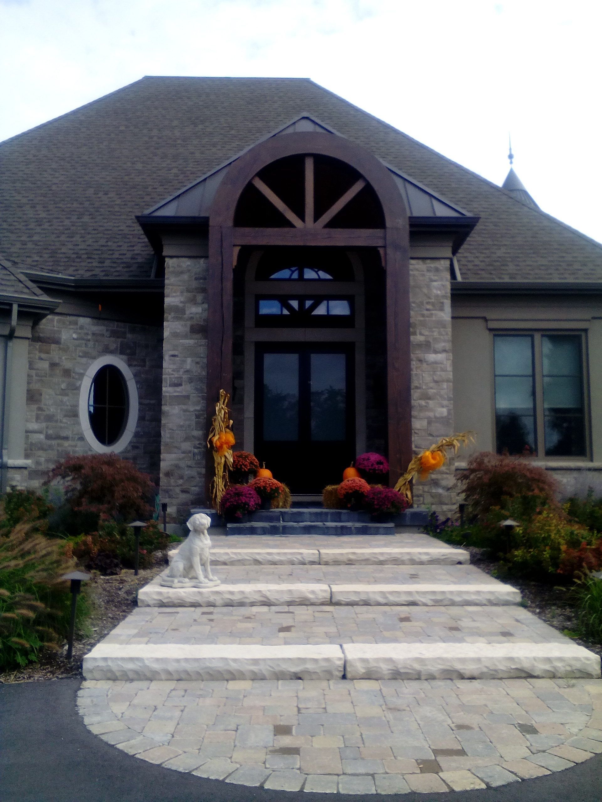 A stone house entrance with an arched wooden porch, a set of wide stone steps, and potted autumn flowers by the door.