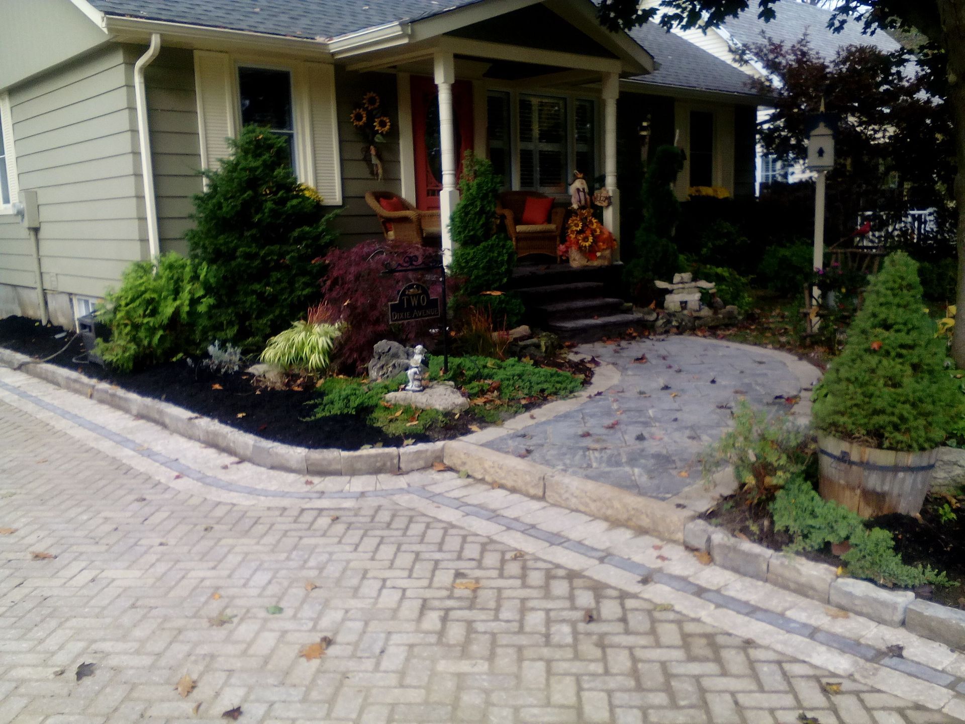 A herringbone-patterned brick driveway leads to a stone walkway, landscaped garden beds, and the porch of a small house.