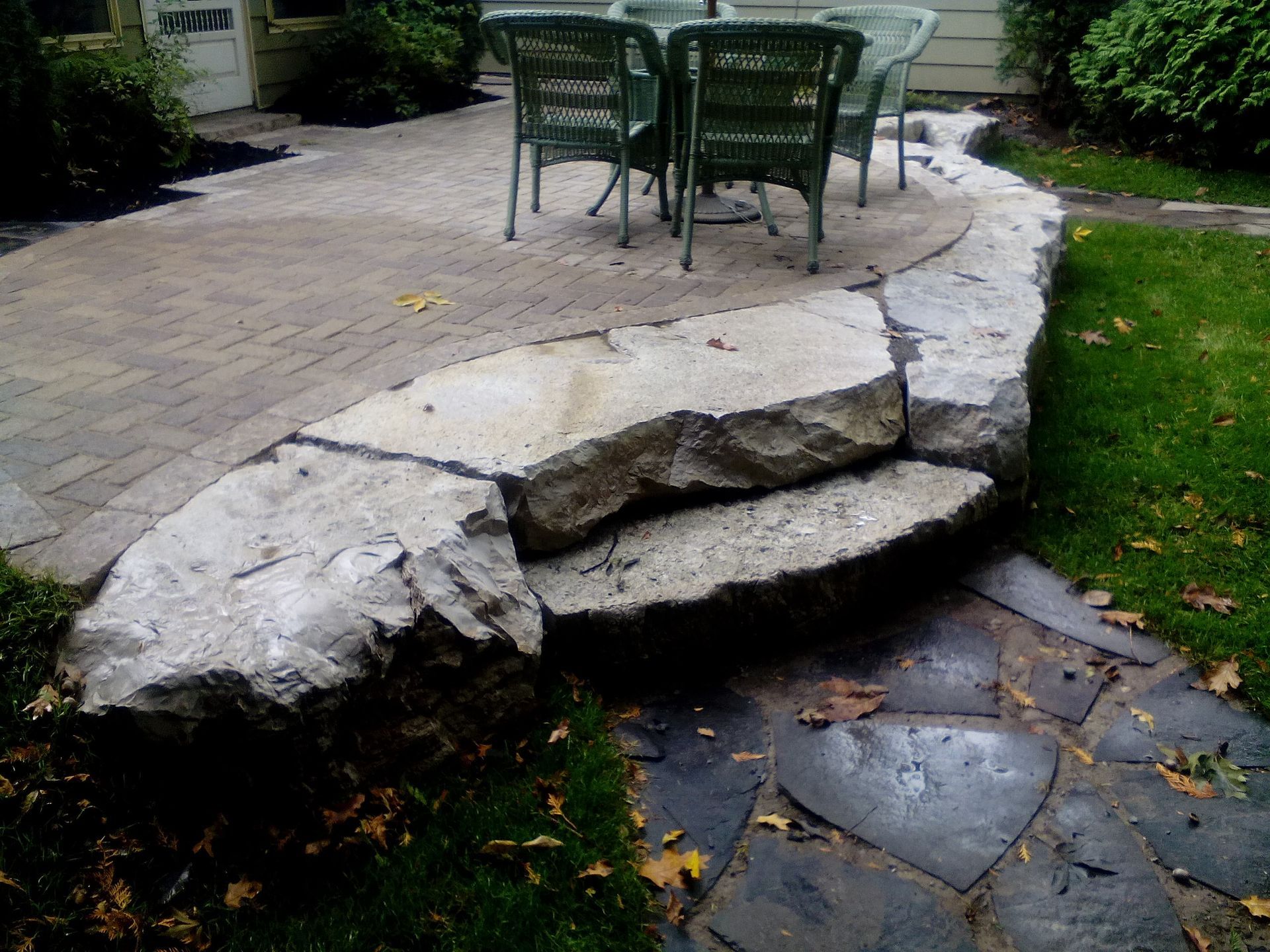 Two large, rough-cut stone steps lead from a lawn path up to a circular paved patio with a bistro table and chairs.
