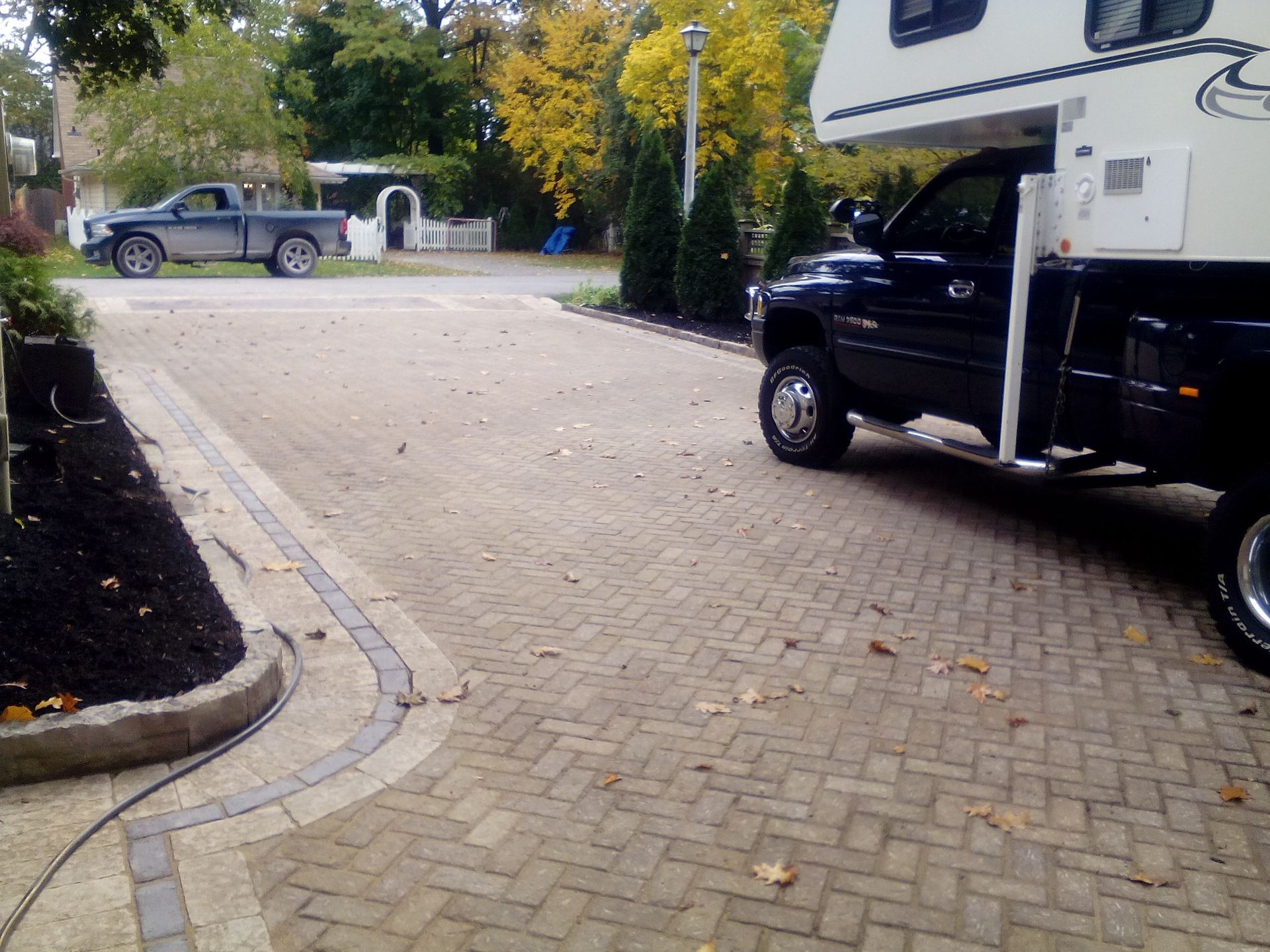 A large truck with a camper unit parked on a herringbone-patterned paved driveway with fallen leaves, near a garden bed.