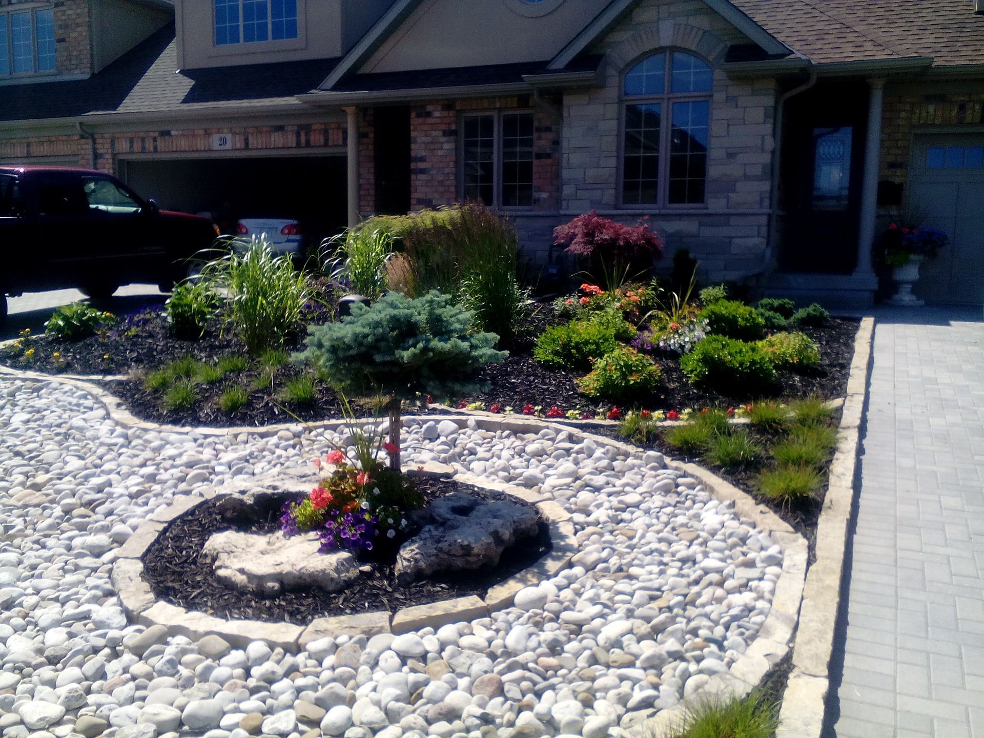 A suburban home exterior with a front yard featuring a circular rock garden, decorative shrubs, and paved landscaping.