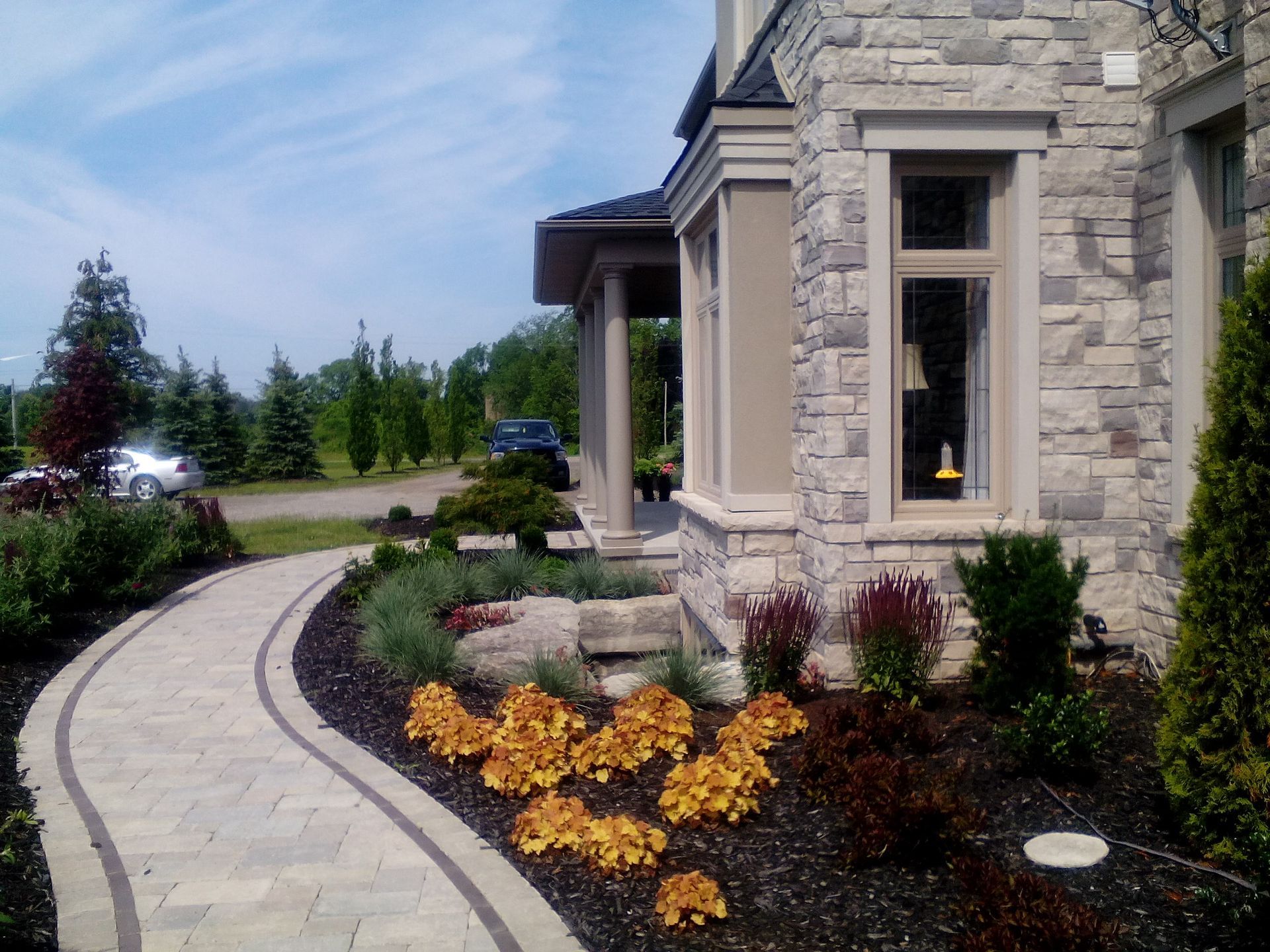 A stone pathway curves through a garden with dark mulch and orange shrubs toward the side of a light-colored stone house.