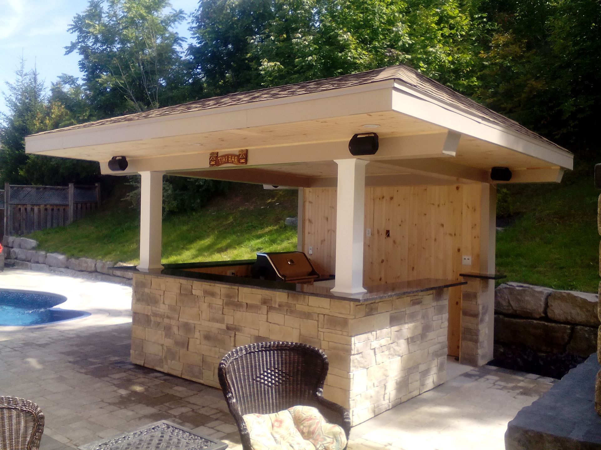 An outdoor stone bar and grill pavilion with a covered roof, positioned next to a swimming pool.