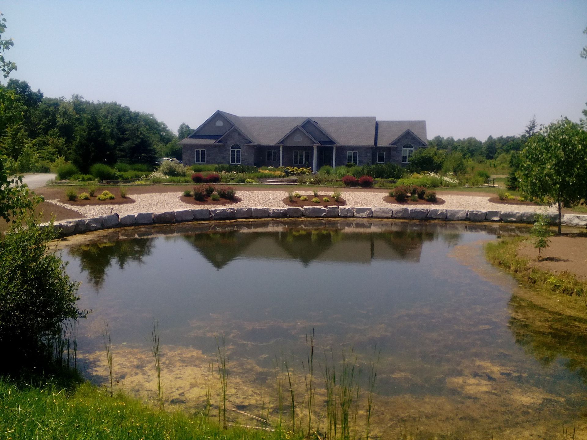 A grey house sits behind a stone-lined pond surrounded by greenery and landscaped garden beds under a clear sky.