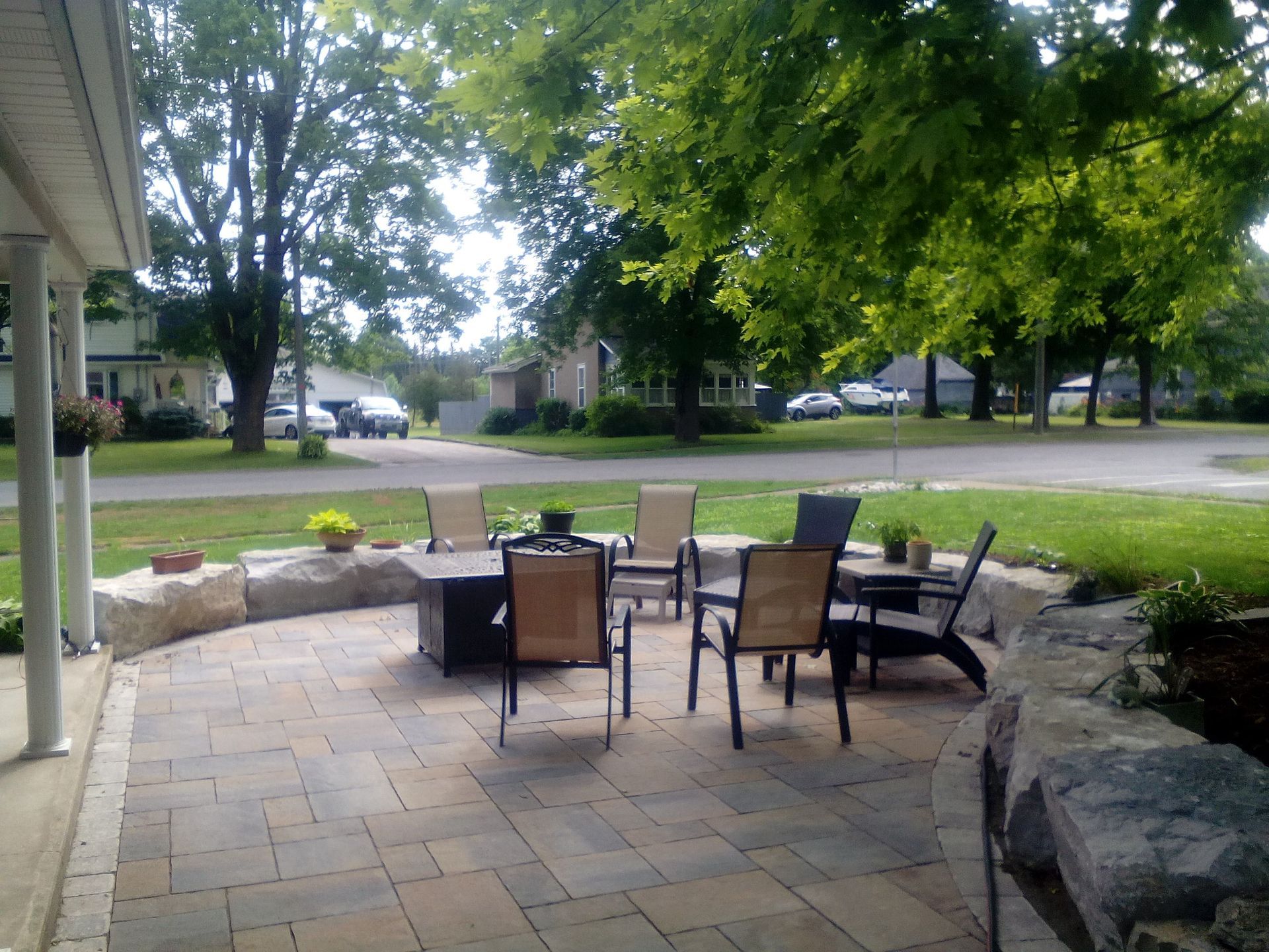 A stone paver patio features chairs arranged around a fire pit, bordered by a rustic stone retaining wall in a yard.