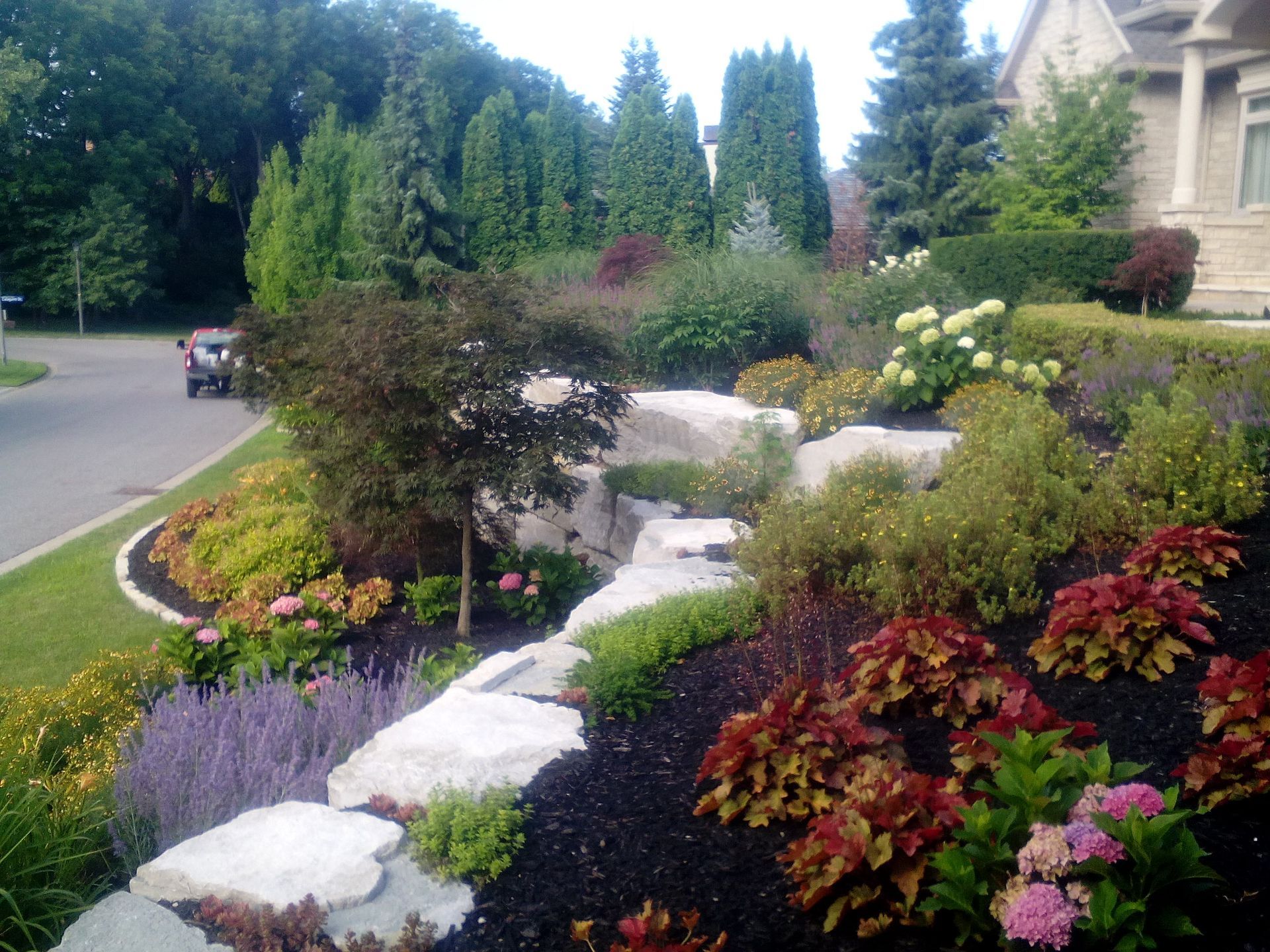 A residential front yard features tiered landscaping with large stones, colorful shrubs, flowers, and dark mulch.