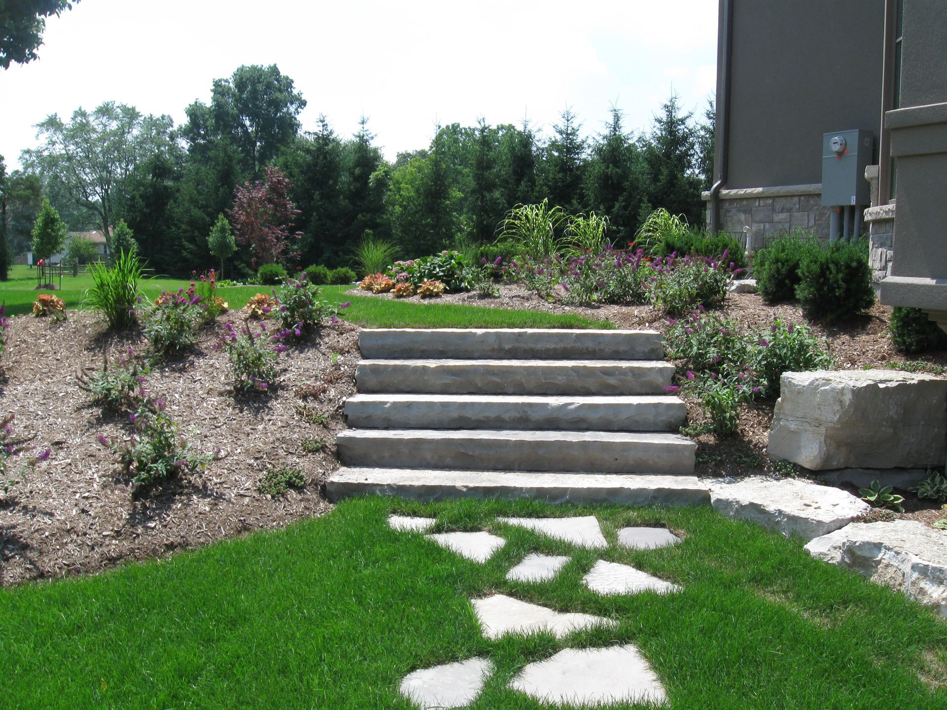Stone steps lead up a grassy hill through a landscaped yard with mulched garden beds and evergreen trees.