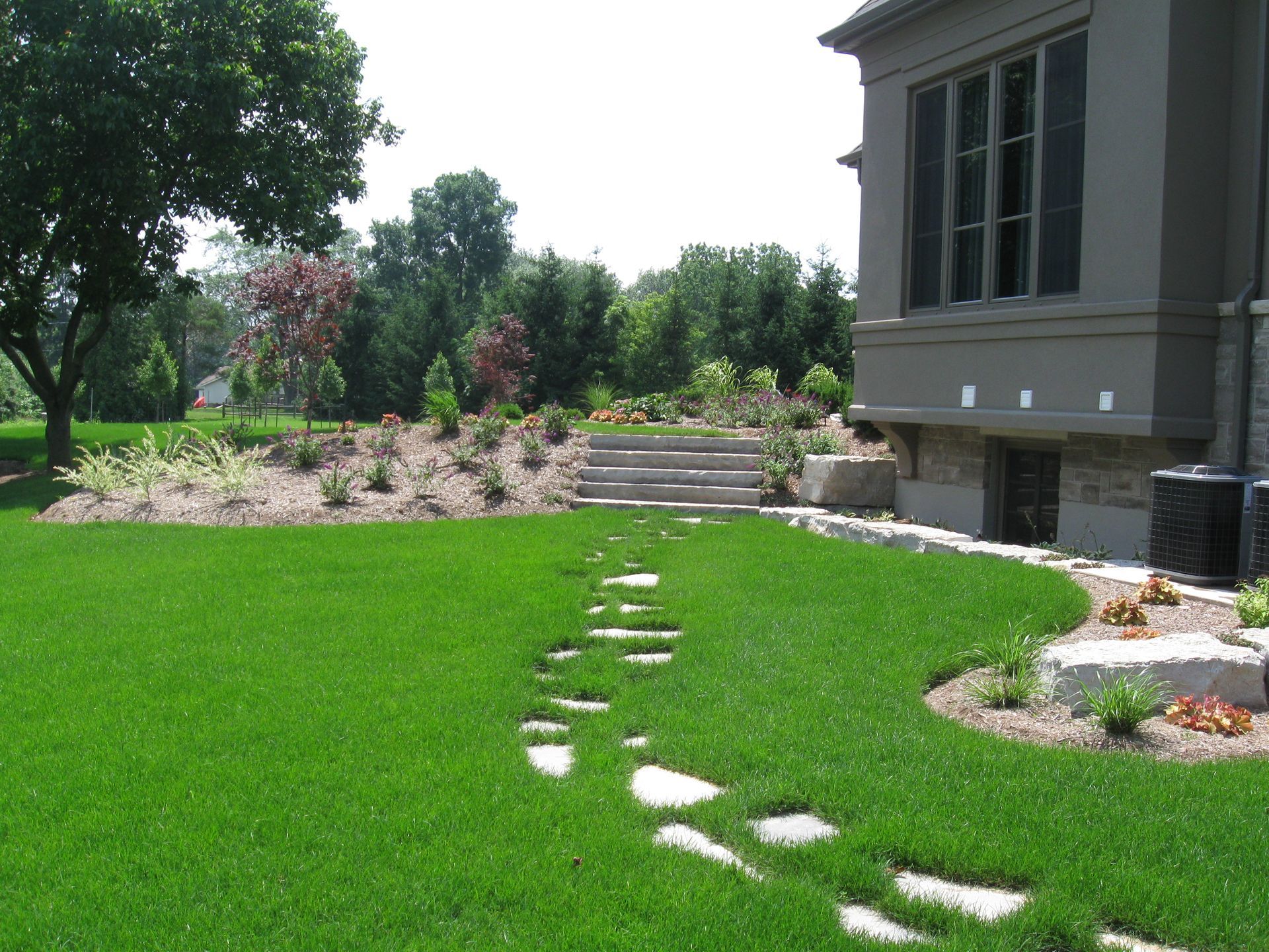 Stone path leading through a lush green lawn to stone steps near a house with brown siding and a large landscaped yard.