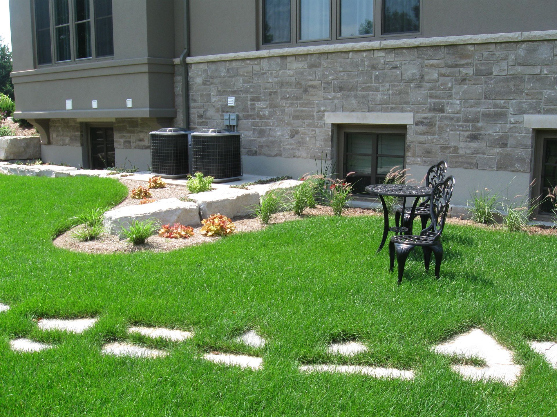 A backyard lawn with a stone path leading to a small patio table and chairs, near a garden bed and a stone house wall.