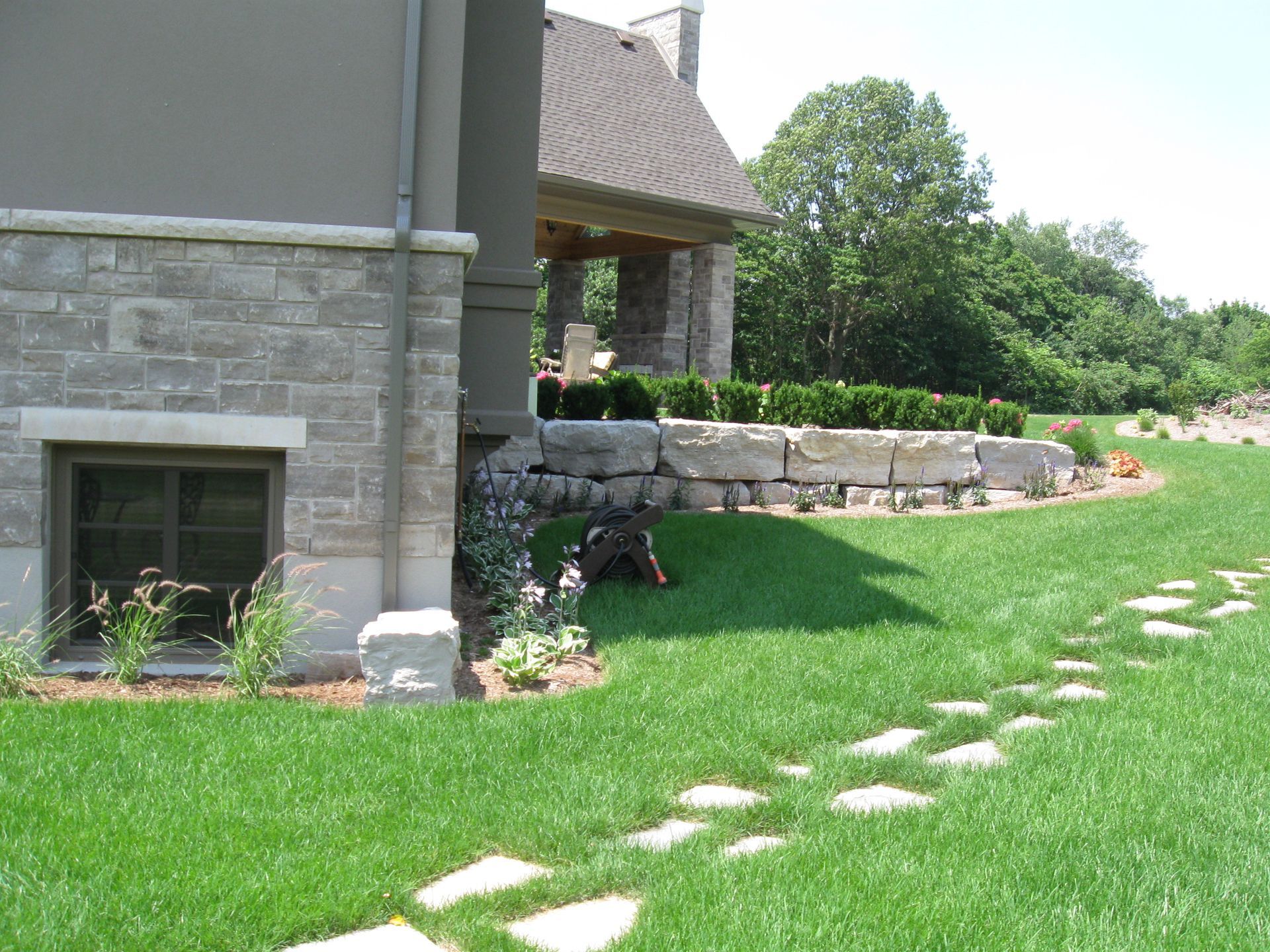 A stone-faced house corner with a basement window, stone retaining wall, and stepping stone path across a lush green lawn.