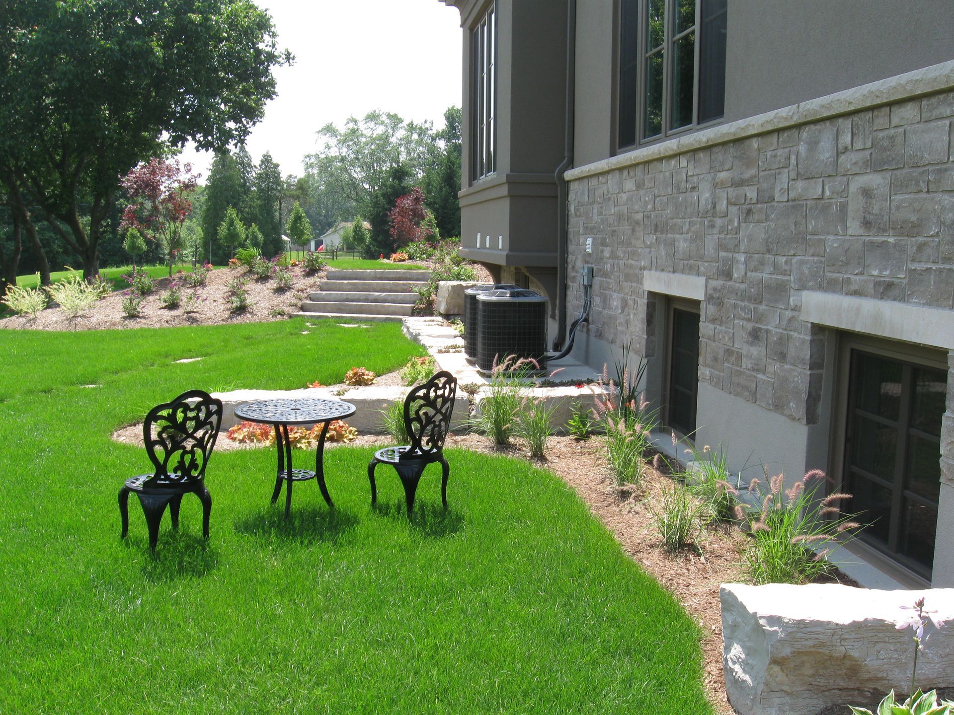 A patio set with a round table and two black metal chairs on a lawn beside a stone-clad house and landscaped garden.