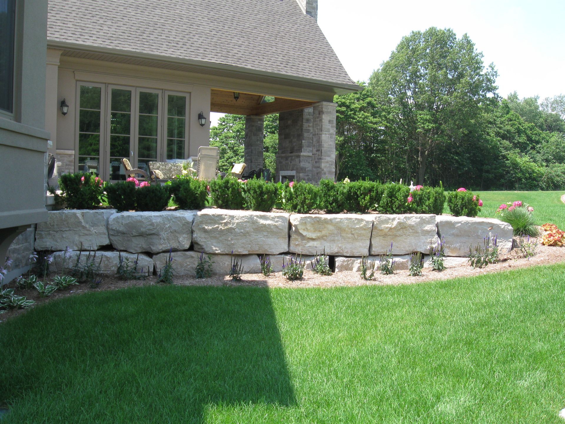 A stone retaining wall sits in front of a house patio, surrounded by a manicured lawn, shrubs, and small green plants.