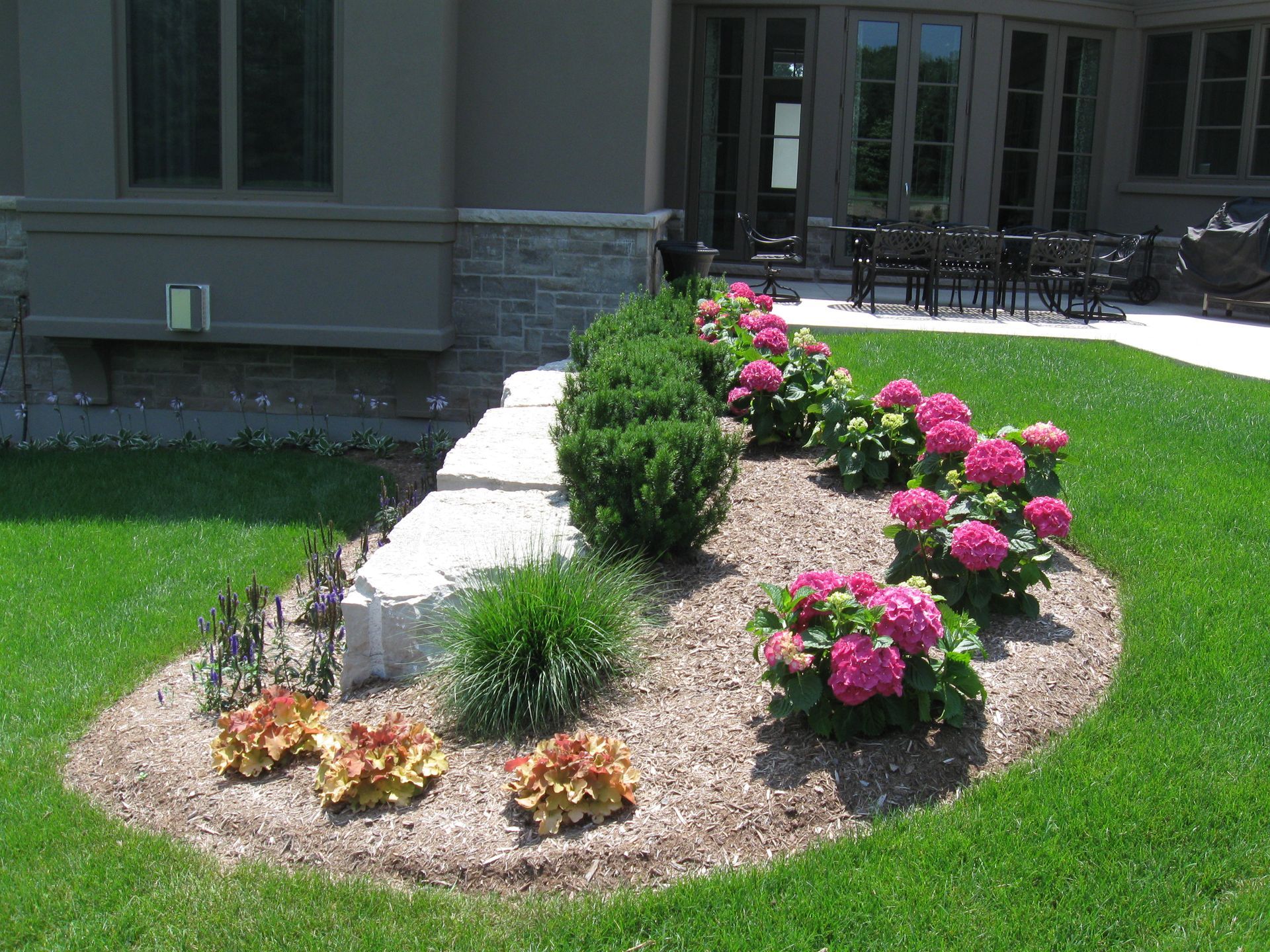 A landscaped garden bed with pink hydrangeas, small green shrubs, and orange plants along a stone retaining wall.