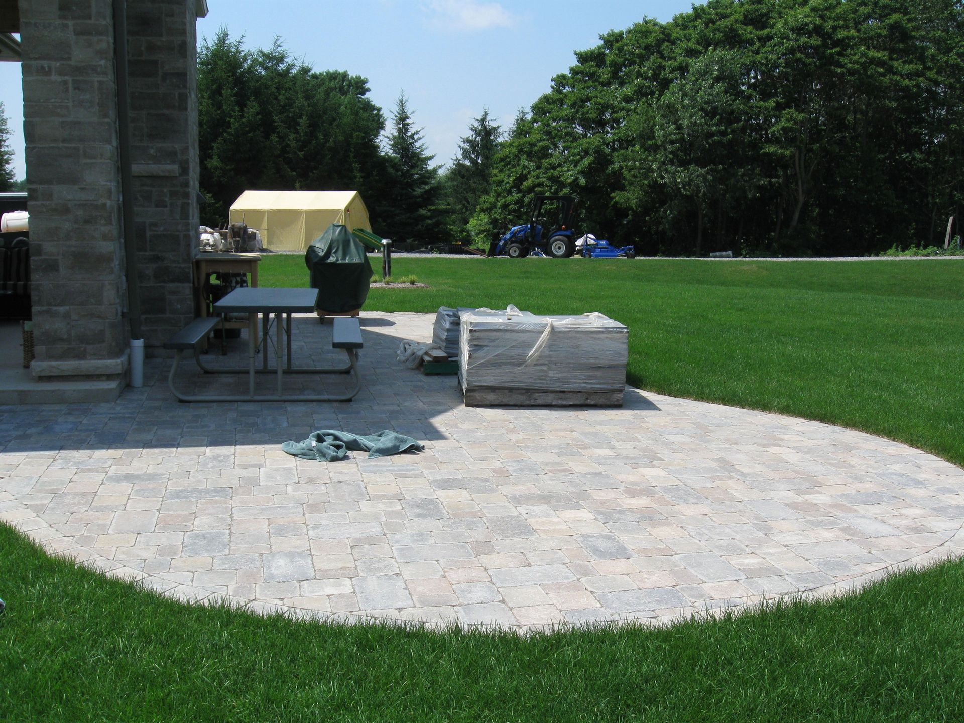A paved stone patio with a picnic table and large stone block sits in a grassy backyard next to a stone pillar structure.