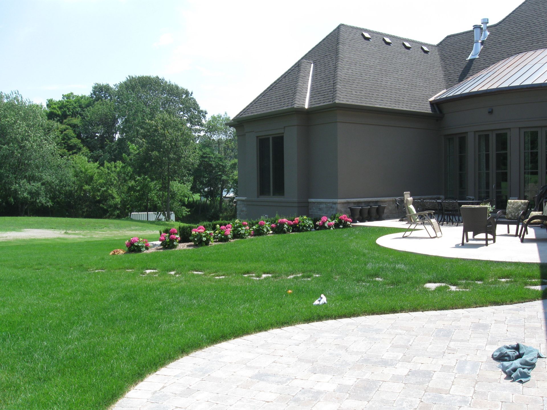 A tan house with a dark roof overlooks a large green lawn, a stone patio, and a row of flowering bushes.