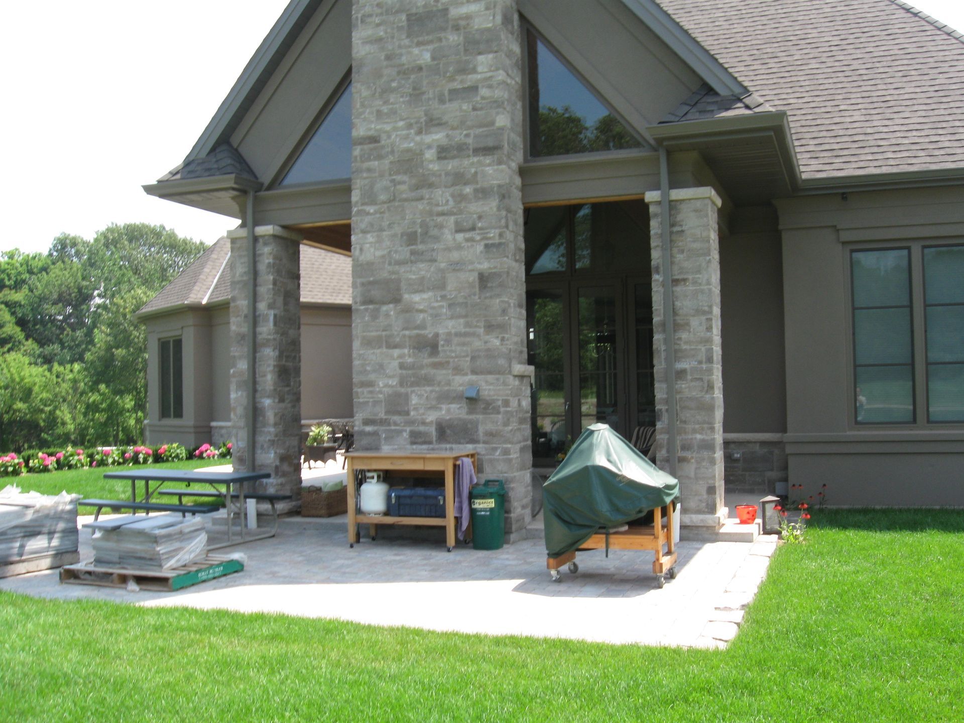 A stone-clad patio with a covered porch, a wooden prep table, and a grill under a green cover, set against a brown house.