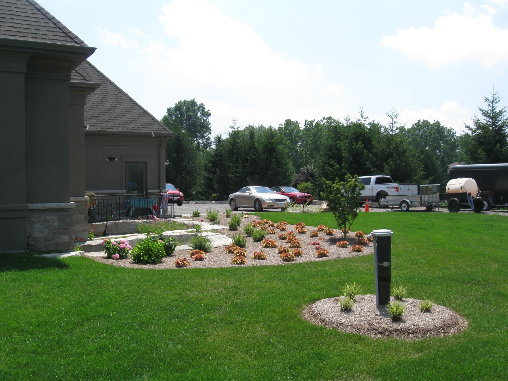 A house exterior with a curved landscaping bed featuring small plants, stone accents, and a grass lawn with a utility post.