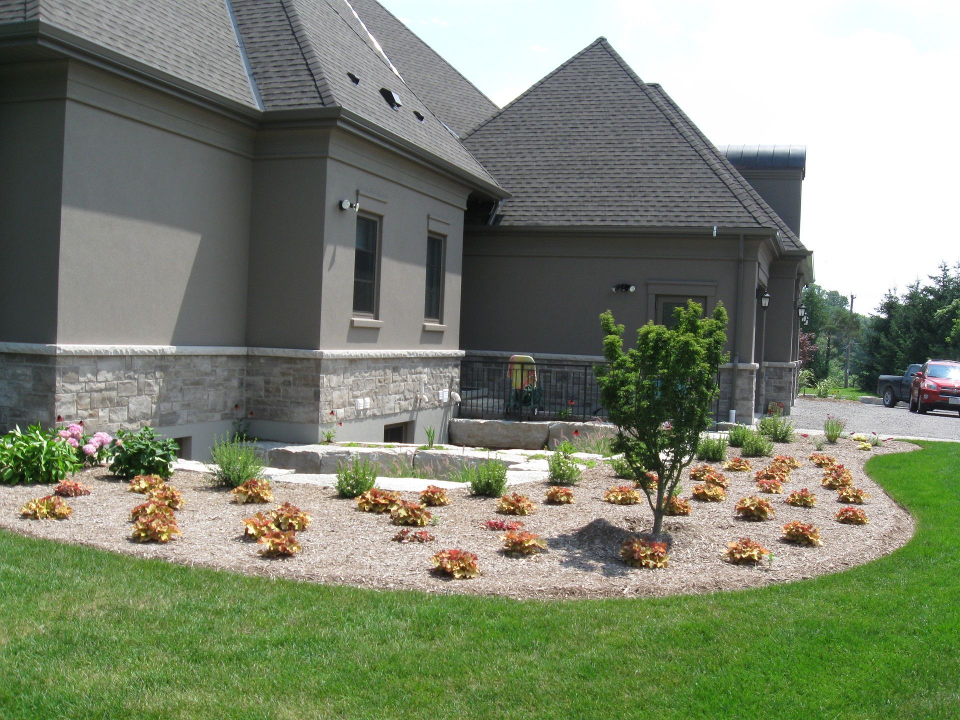 A tan building with a stone foundation and dark roof overlooks a landscaped garden bed with mulch, shrubs, and flowers.