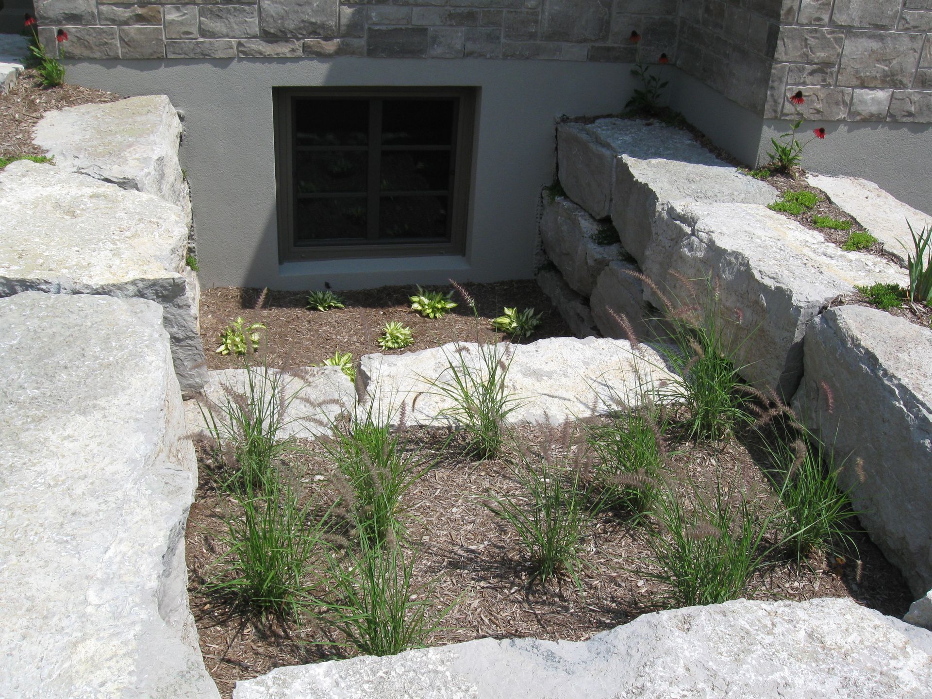 A basement window set into a stone foundation, surrounded by a tiered stone retaining wall and a landscaped garden bed.