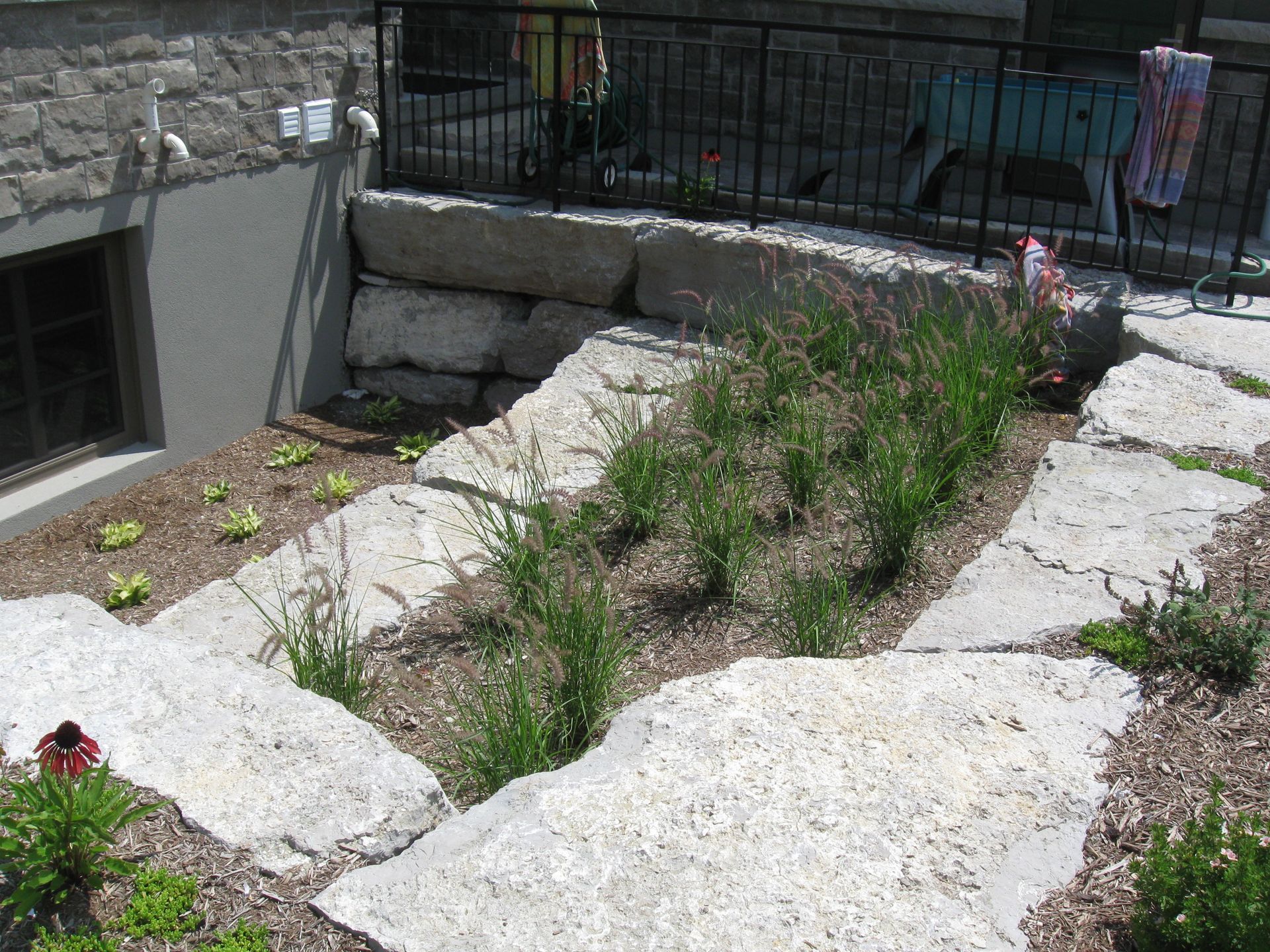 Tiered landscape garden featuring large grey limestone slabs and various green plants next to a house foundation.