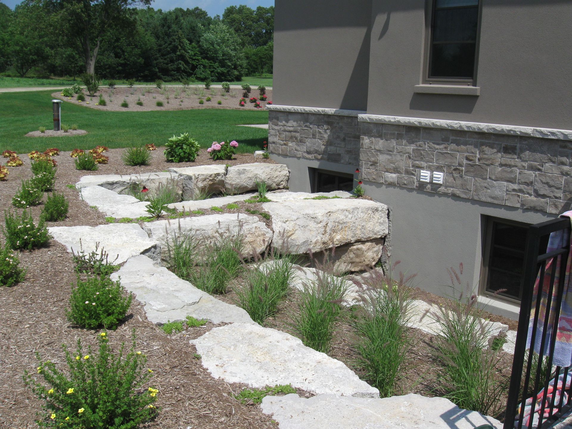 A stone-retained garden bed with small plants slopes downward toward the basement windows of a grey stone house.
