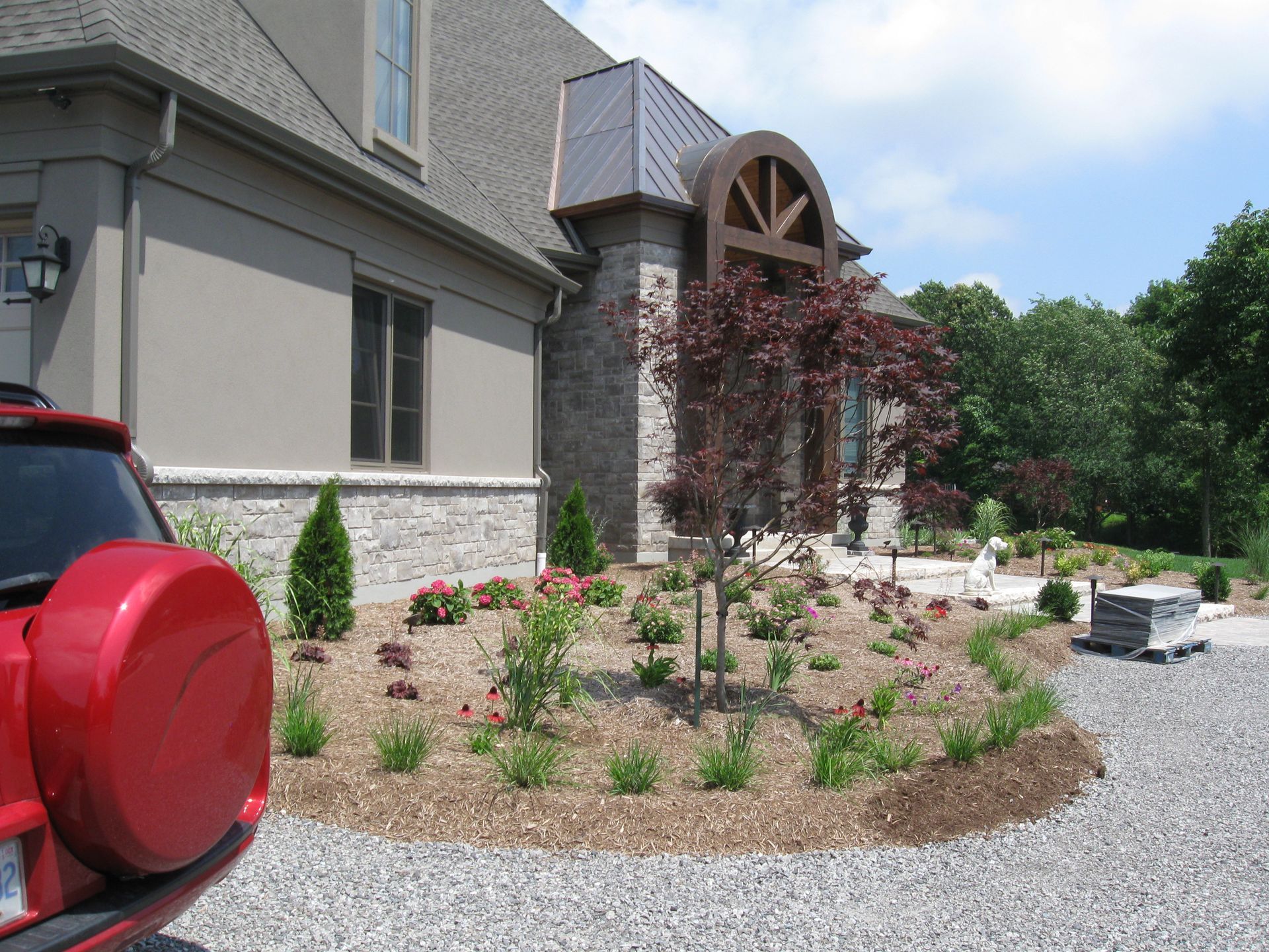 A tan home with stone accents and a curved wooden entryway, featuring a front garden with mulch and a red vehicle parked.