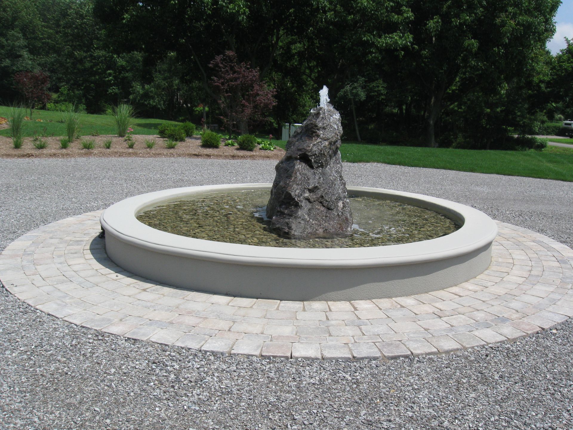 A rock fountain with a water spout sits in a circular, paved basin surrounded by a gravel driveway and green landscaping.