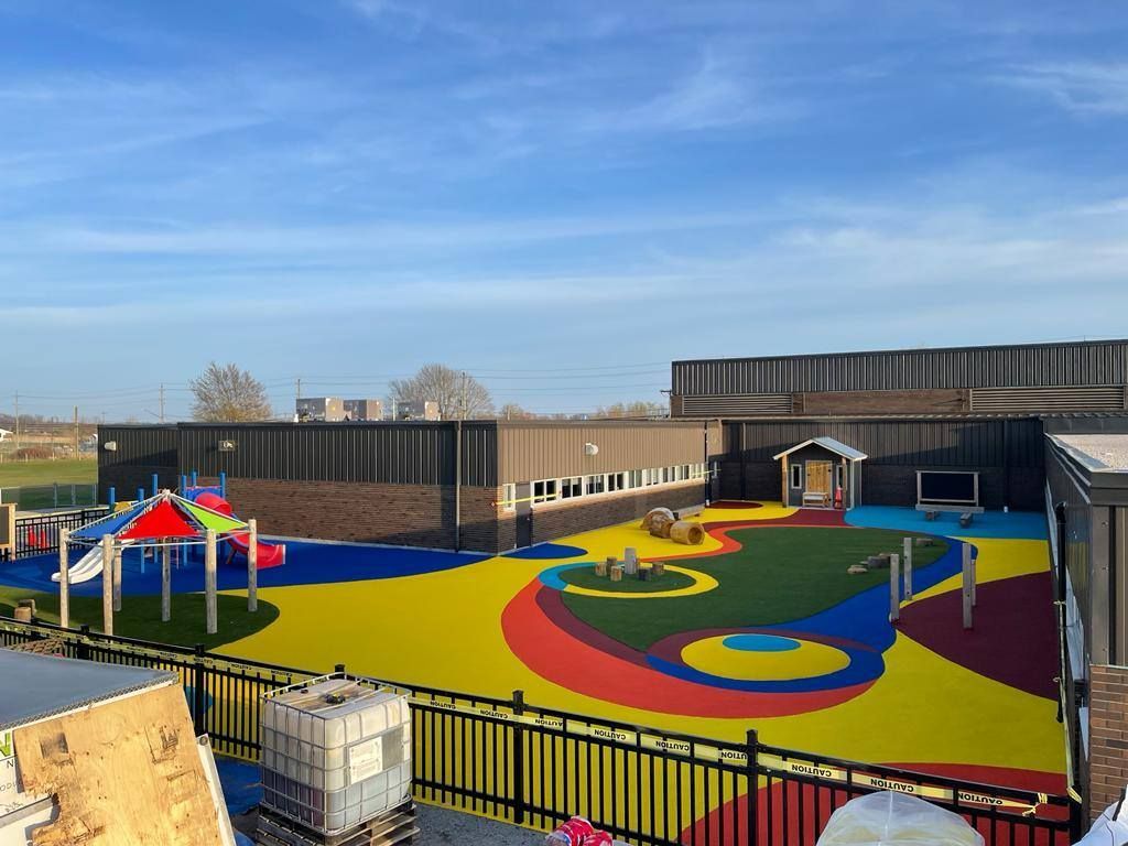 A playground with a colorful, abstract-patterned rubber surface, a small climbing structure, and a fenced-in play area.